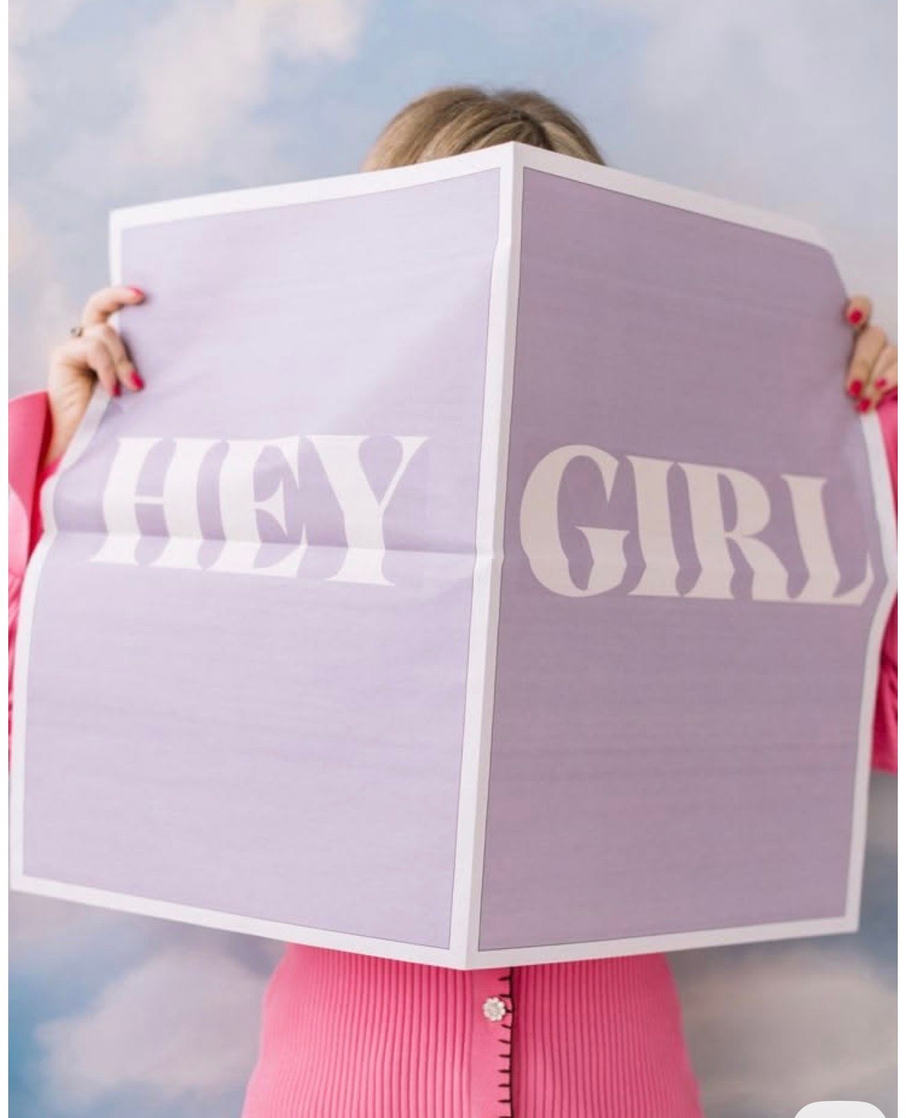 Person holding a large lavender poster with bold text reading HEY GIRL against a blue sky background.