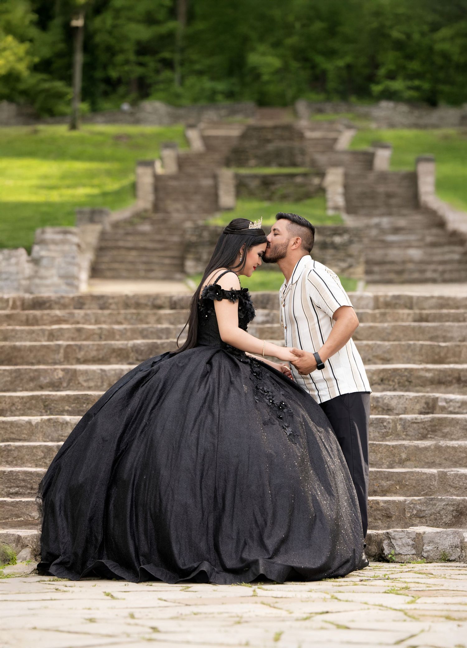 Couple sharing a romantic kiss outdoors, woman in elegant black ball gown, man in striped shirt, stone steps background.
