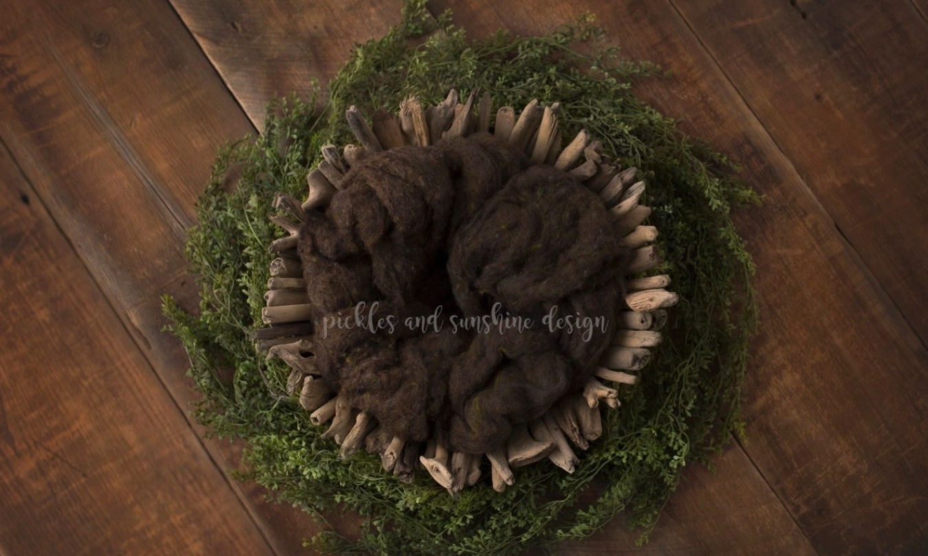 Dark brown fur nest surrounded by green moss wreath on wooden floor for newborn photography.