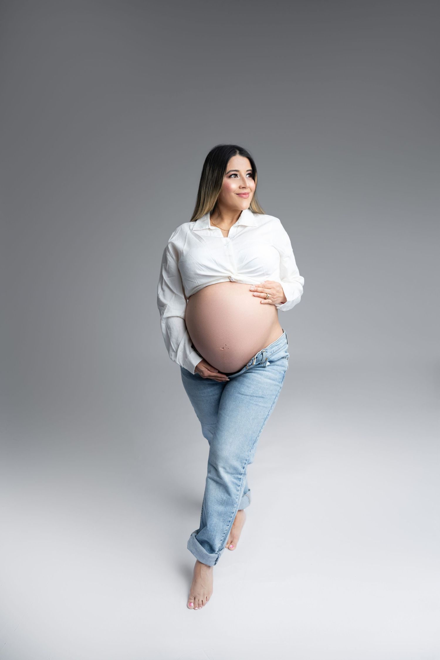 Pregnant woman in white shirt and jeans poses barefoot against gray background in maternity photoshoot.
