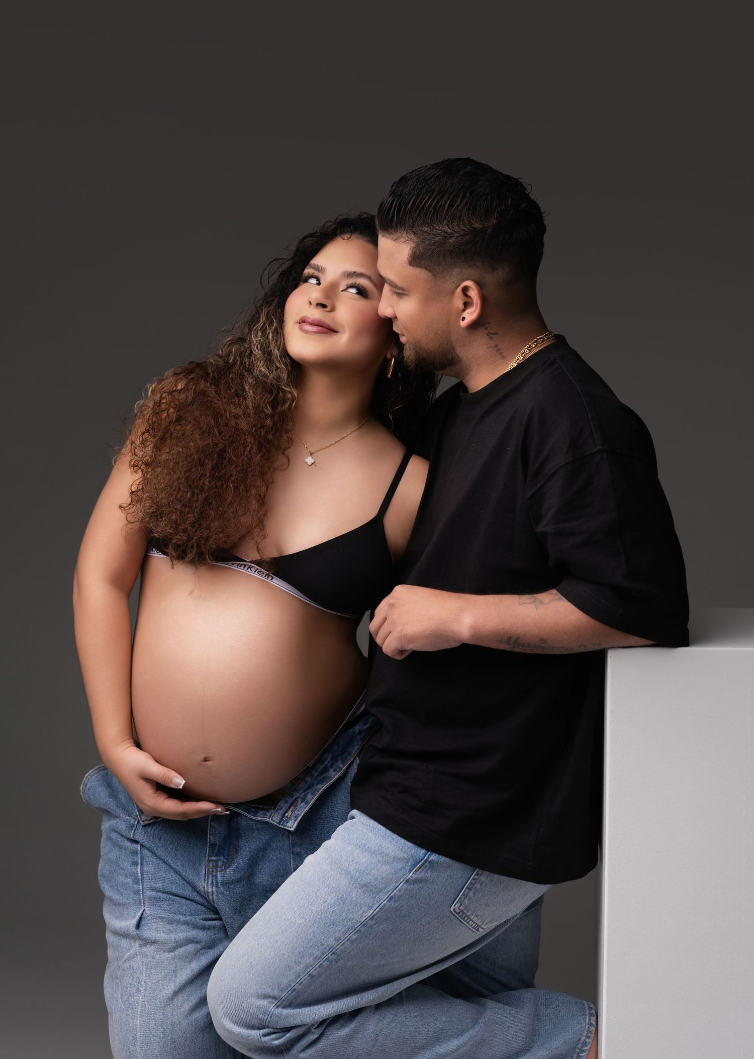 A couple in black and denim embrace tenderly during a maternity photoshoot against a gray studio backdrop.