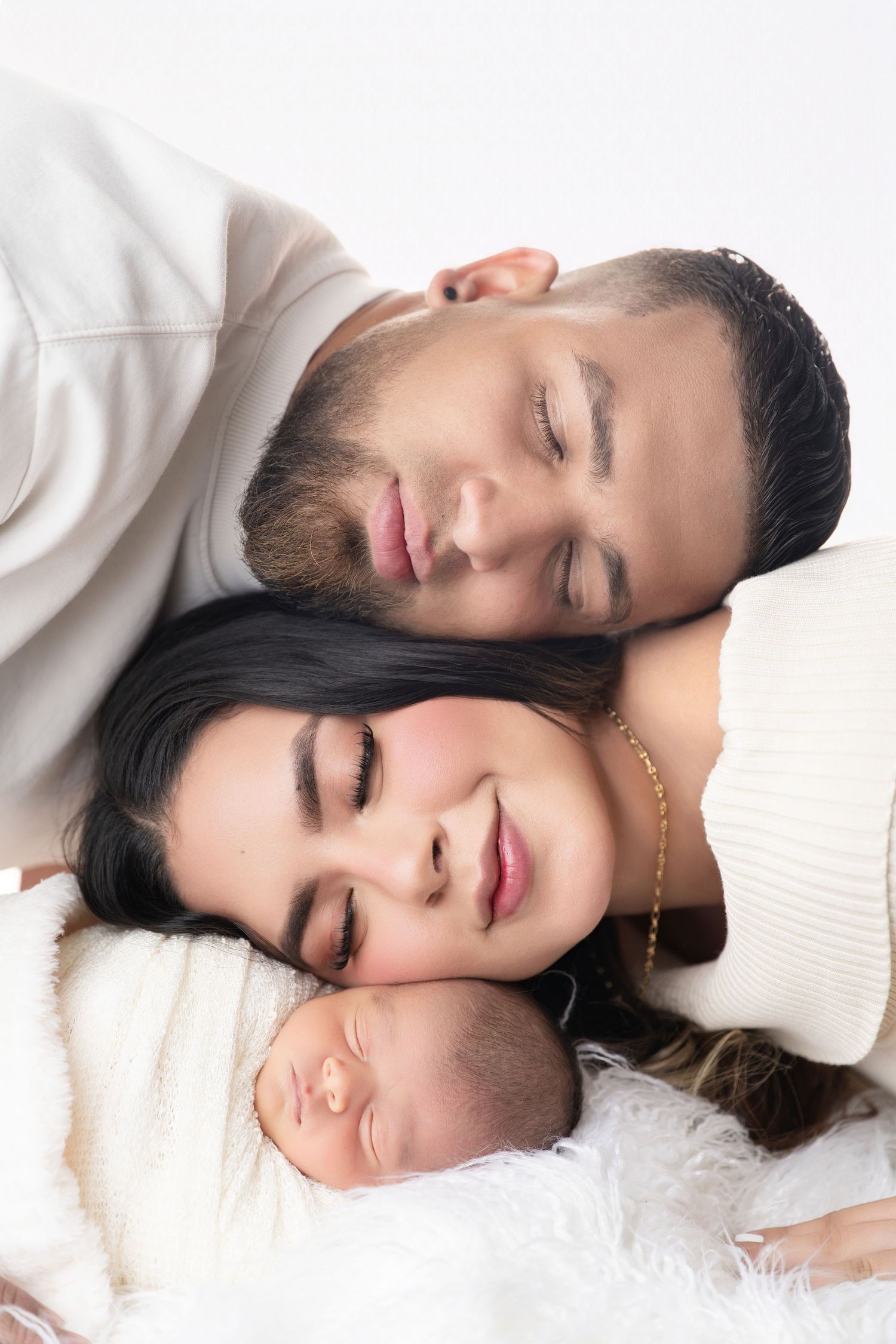 A couple with closed eyes resting their heads near their sleeping newborn baby on a white fluffy blanket.
