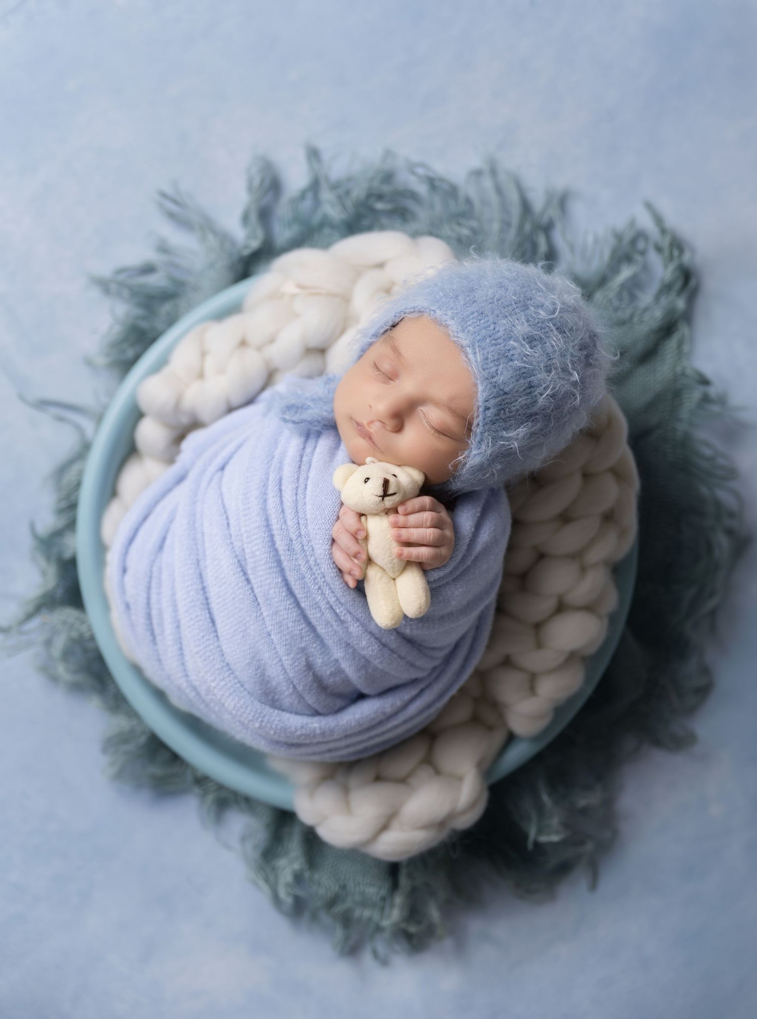 Newborn baby wrapped in blue swaddle holding tiny teddy bear, sleeping in bowl with white knit blanket and blue fur.