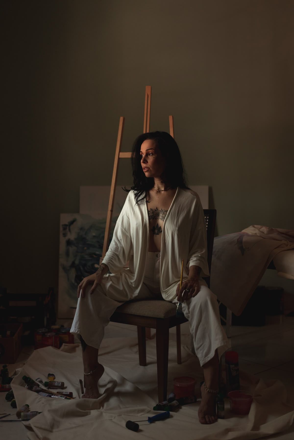 A tattooed female artist sits on a wooden chair in a dimly lit studio with an easel, canvases, and paint supplies.