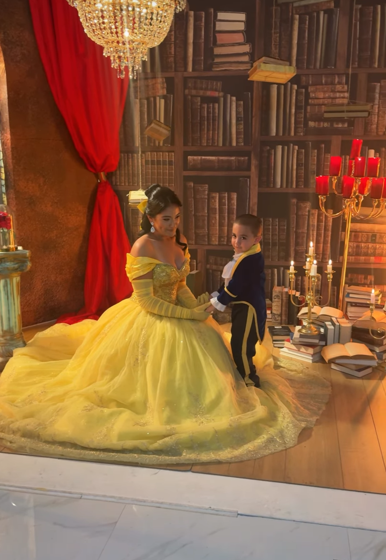 Woman in yellow Belle gown dances with child in Beast costume in enchanted library setting with red curtains and candelabras.