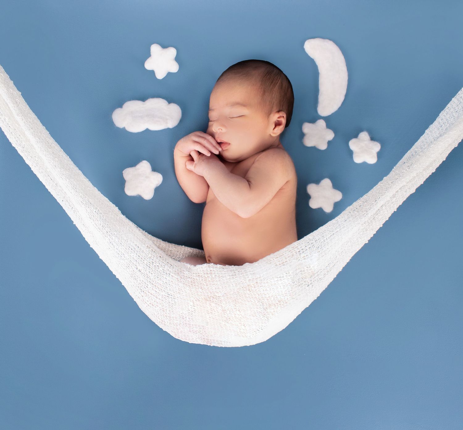 Newborn baby sleeping peacefully in a white hammock on blue background with cloud and star decorations.