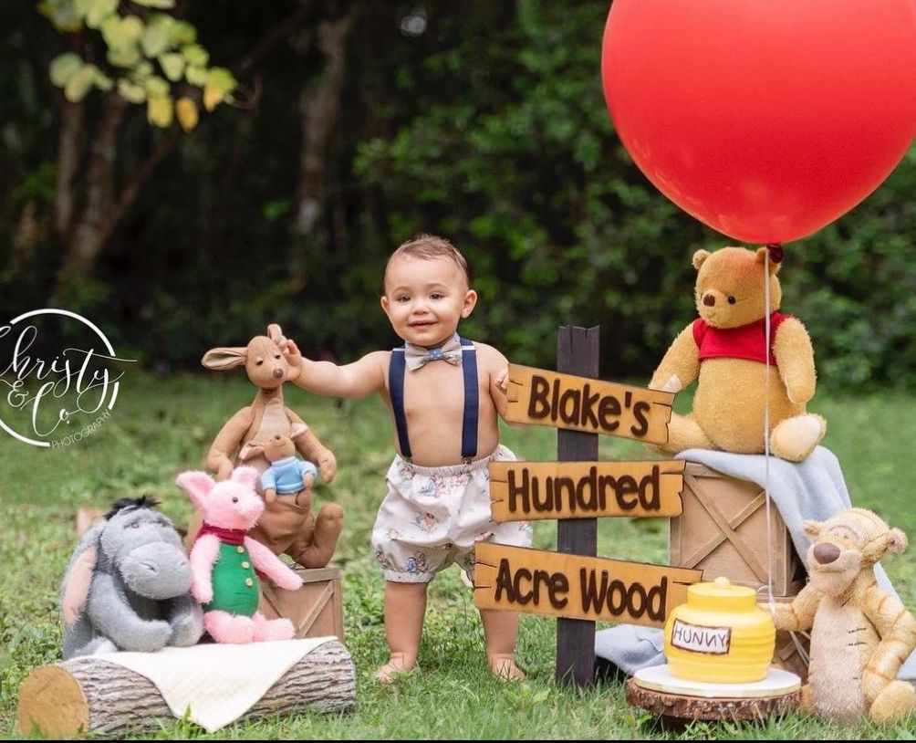 Baby boy celebrates first birthday with Winnie the Pooh theme, surrounded by stuffed animals and a red balloon outdoors.