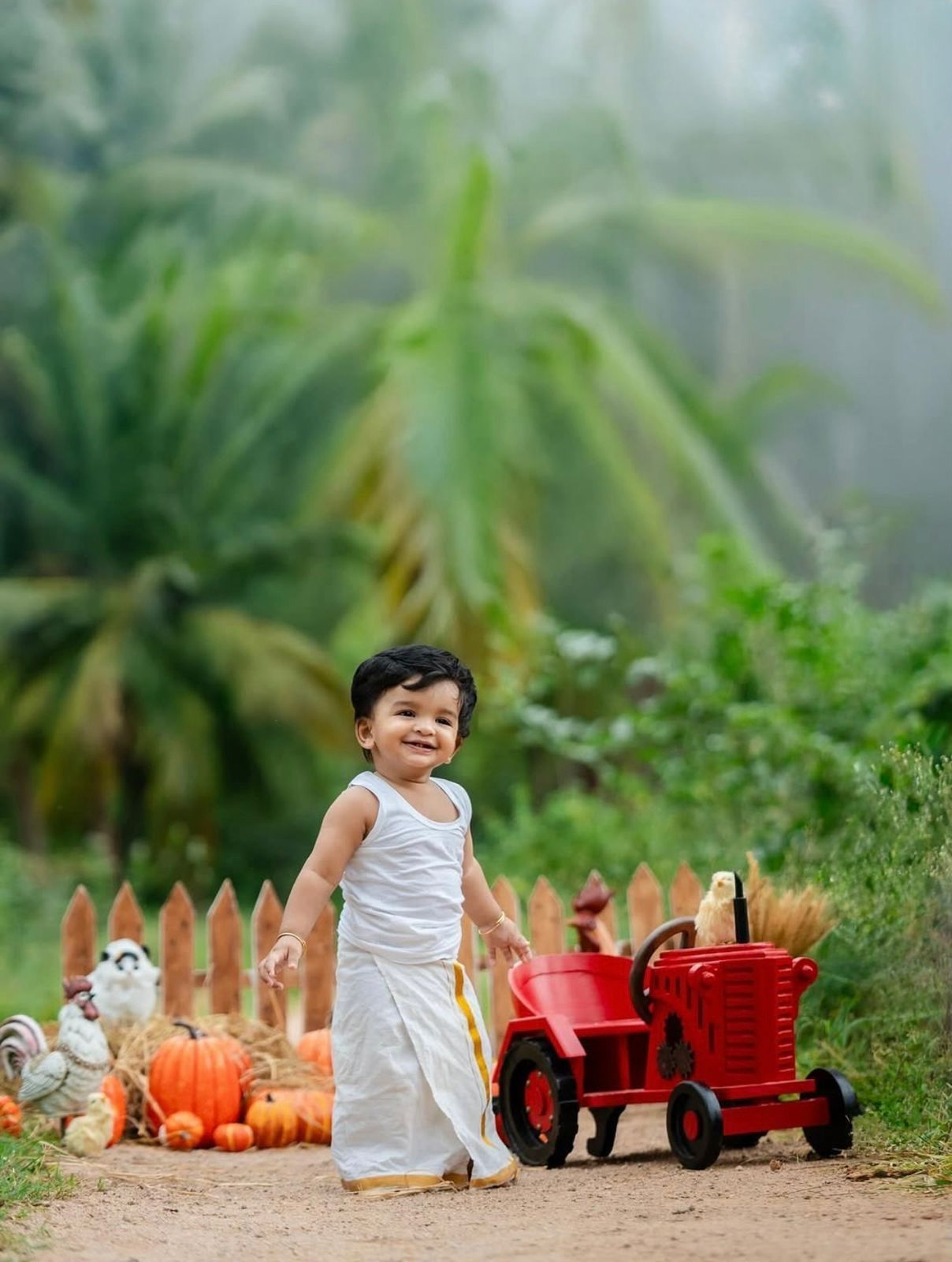 Smiling toddler boy in white outfit standing beside a red toy tractor with pumpkins and a rooster outdoors.