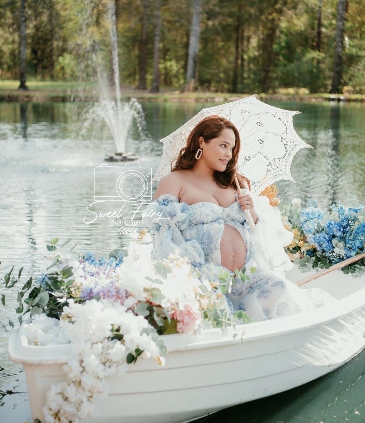 Pregnant woman in blue gown holding white parasol on flower-adorned boat on serene lake with fountain.
