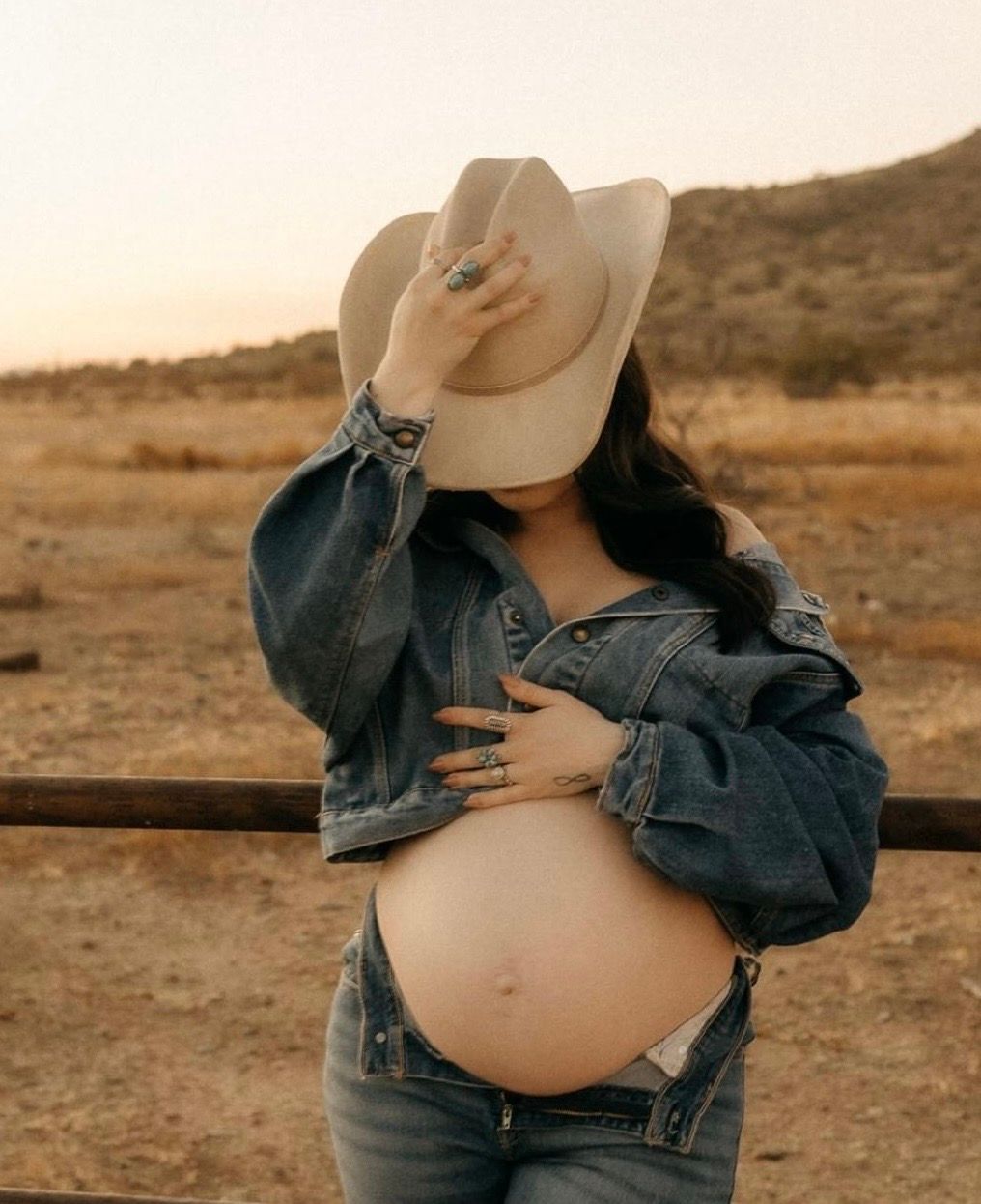 Pregnant woman in denim jacket and cowboy hat posing outdoors in desert landscape at golden hour.