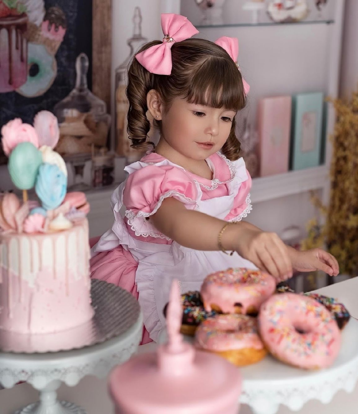 A pink and white themed dessert party setup with decorated cakes, donuts and ruffled details on display.