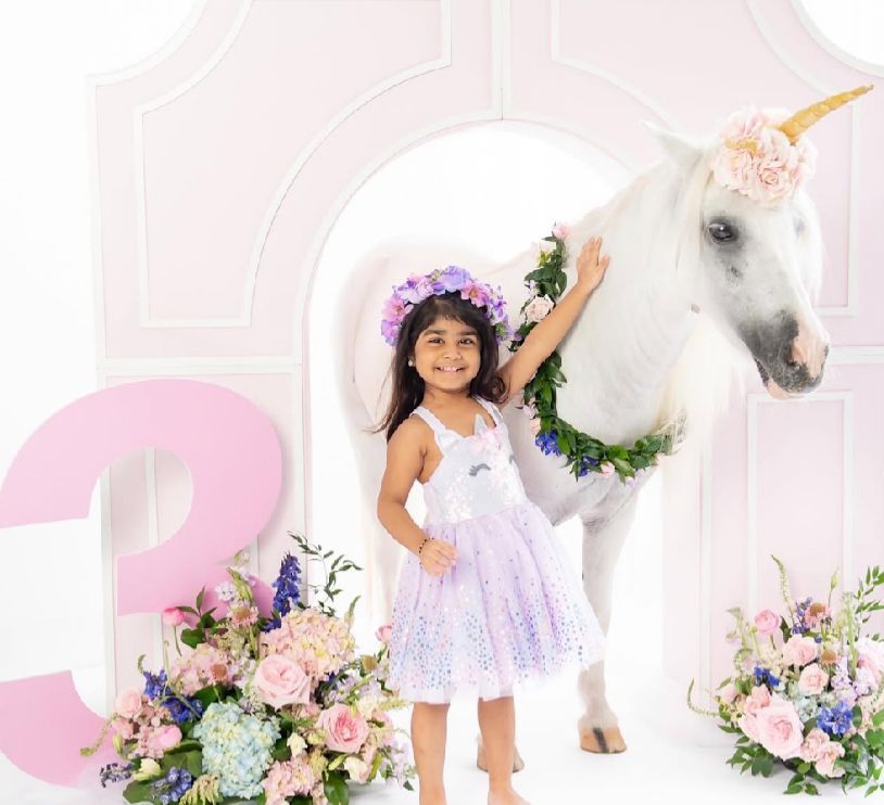 A magical unicorn themed photo session features a child in a lavender dress with floral decorations and pink backdrop.