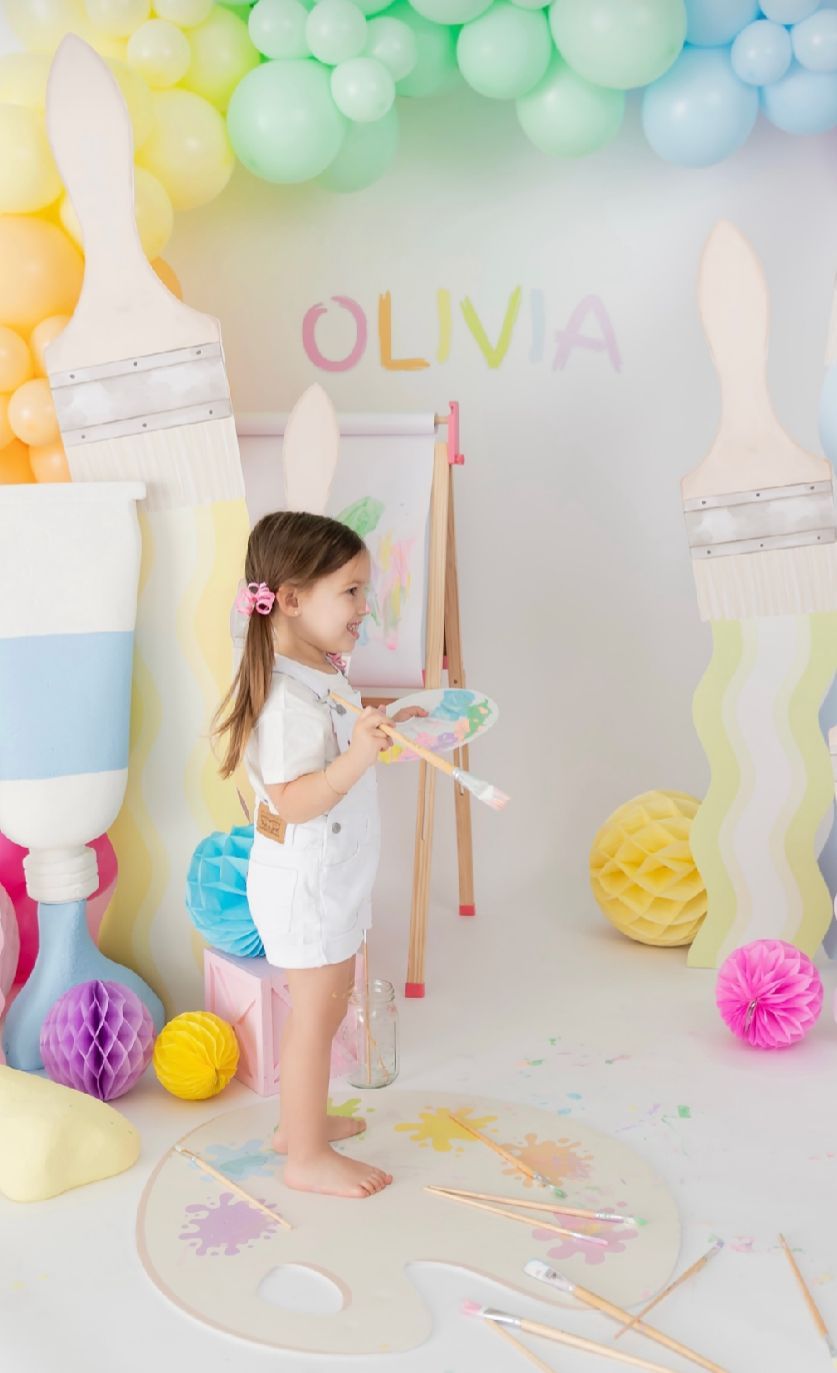 Pastel balloon garland decorates a party setup with colorful paper decorations and art supplies against a white backdrop.
