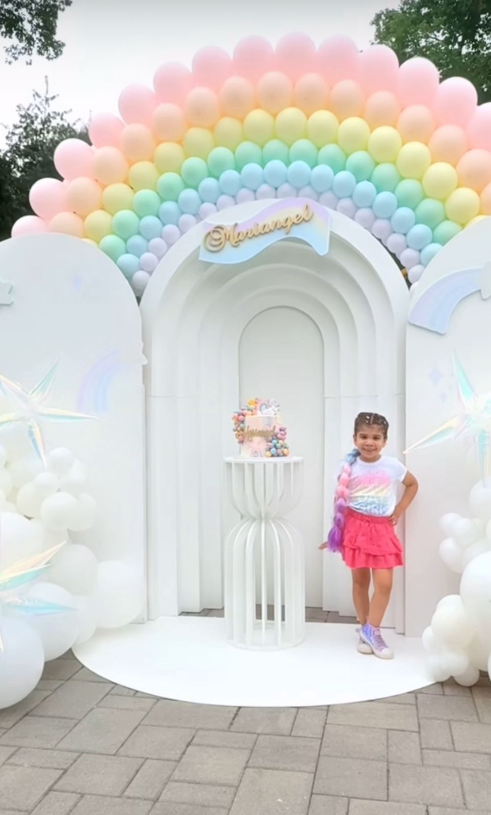 Pastel rainbow balloon arch over white archway with decorative stand and cake display for a party celebration setup.