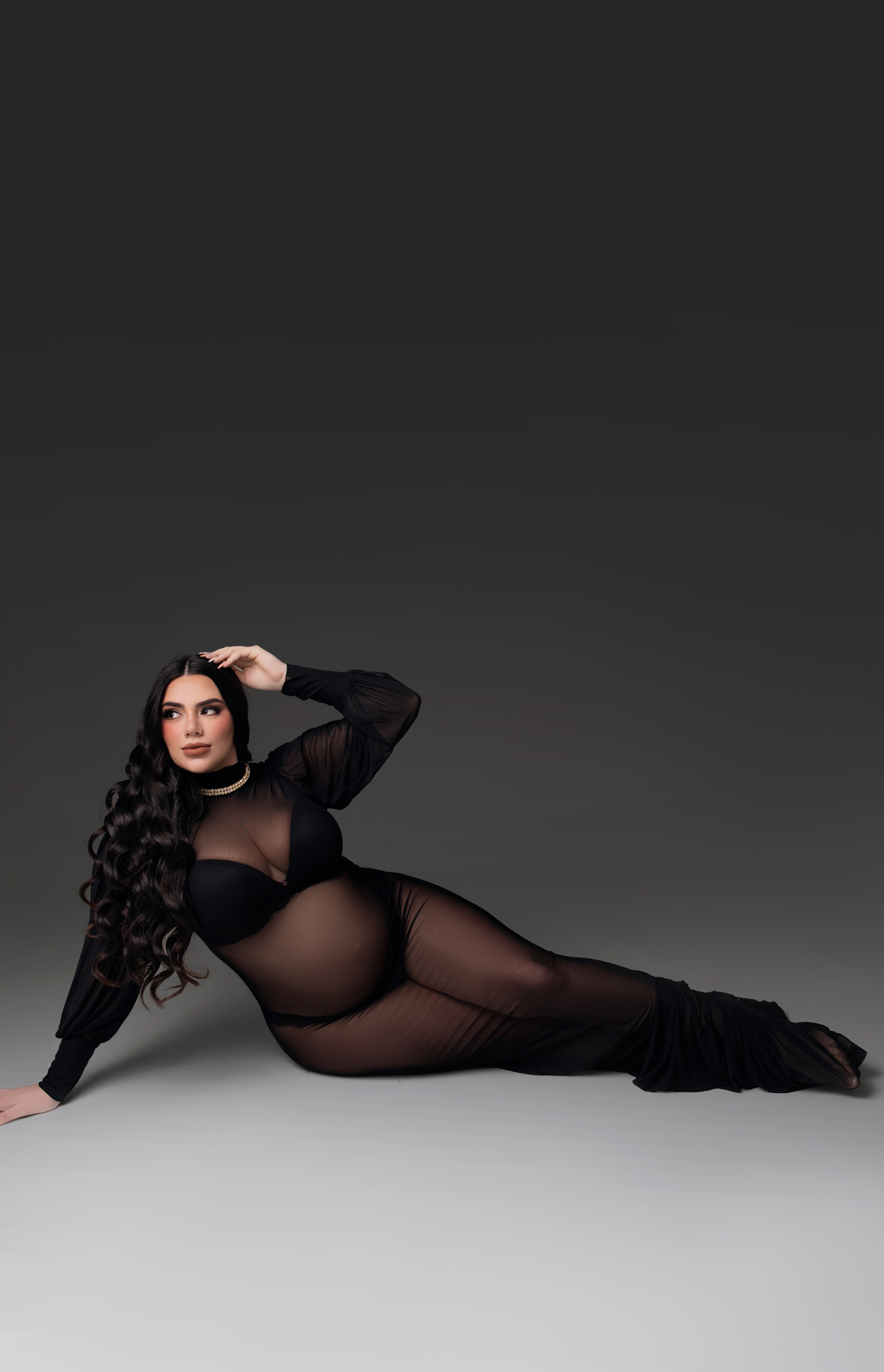 Woman in black sheer long-sleeve gown posing elegantly on floor against dark gray studio background.