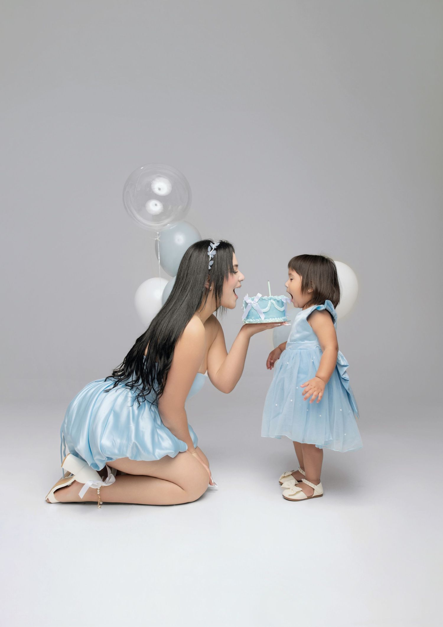 Woman in blue dress presents birthday cake to toddler girl in matching blue dress, surrounded by balloons.