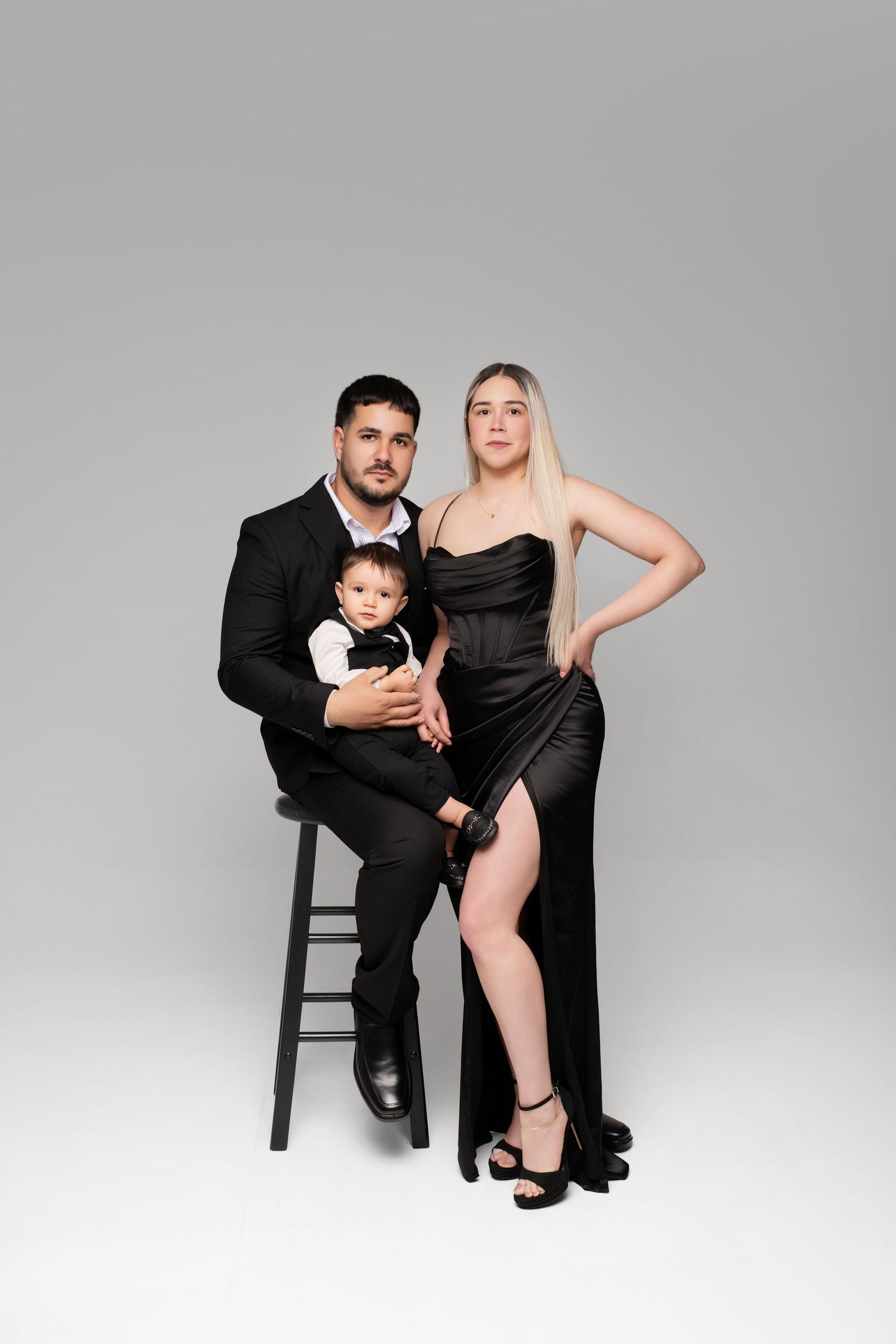 Elegant family portrait with man in black suit holding baby, woman in black satin gown, posing on gray background.