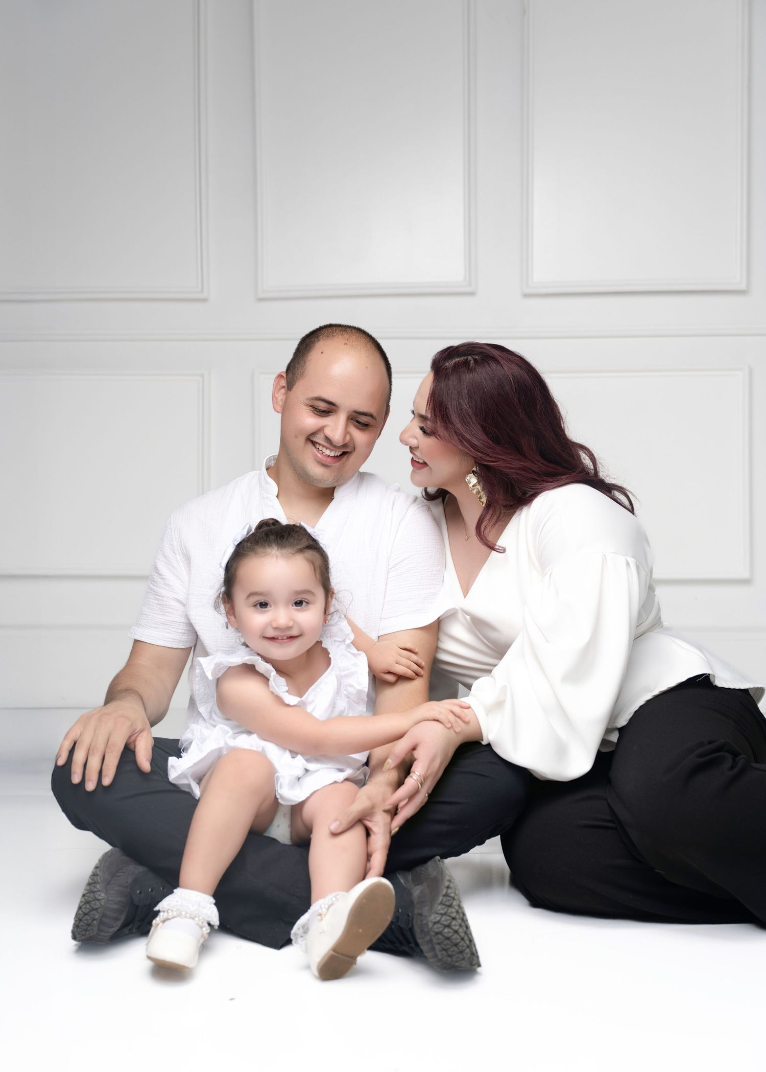 A happy family of three poses in a white studio, with parents smiling at their toddler daughter in a white dress.