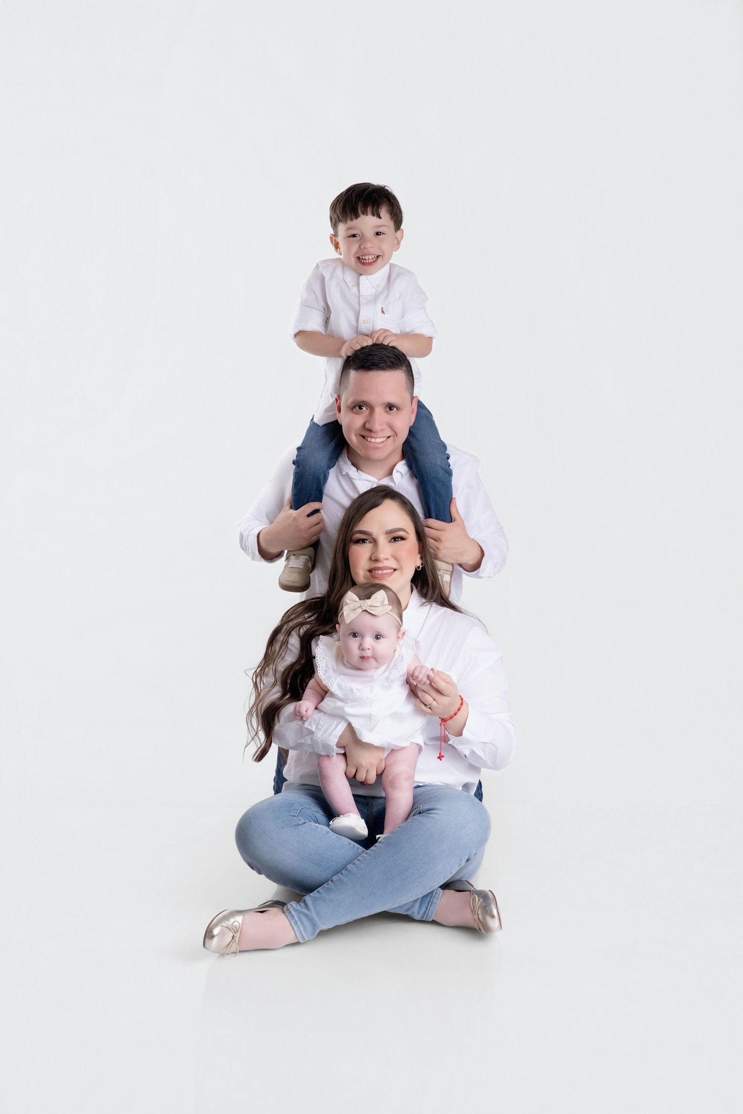 Happy family of four in white shirts and jeans posing together on white background, parents with toddler and baby girl.