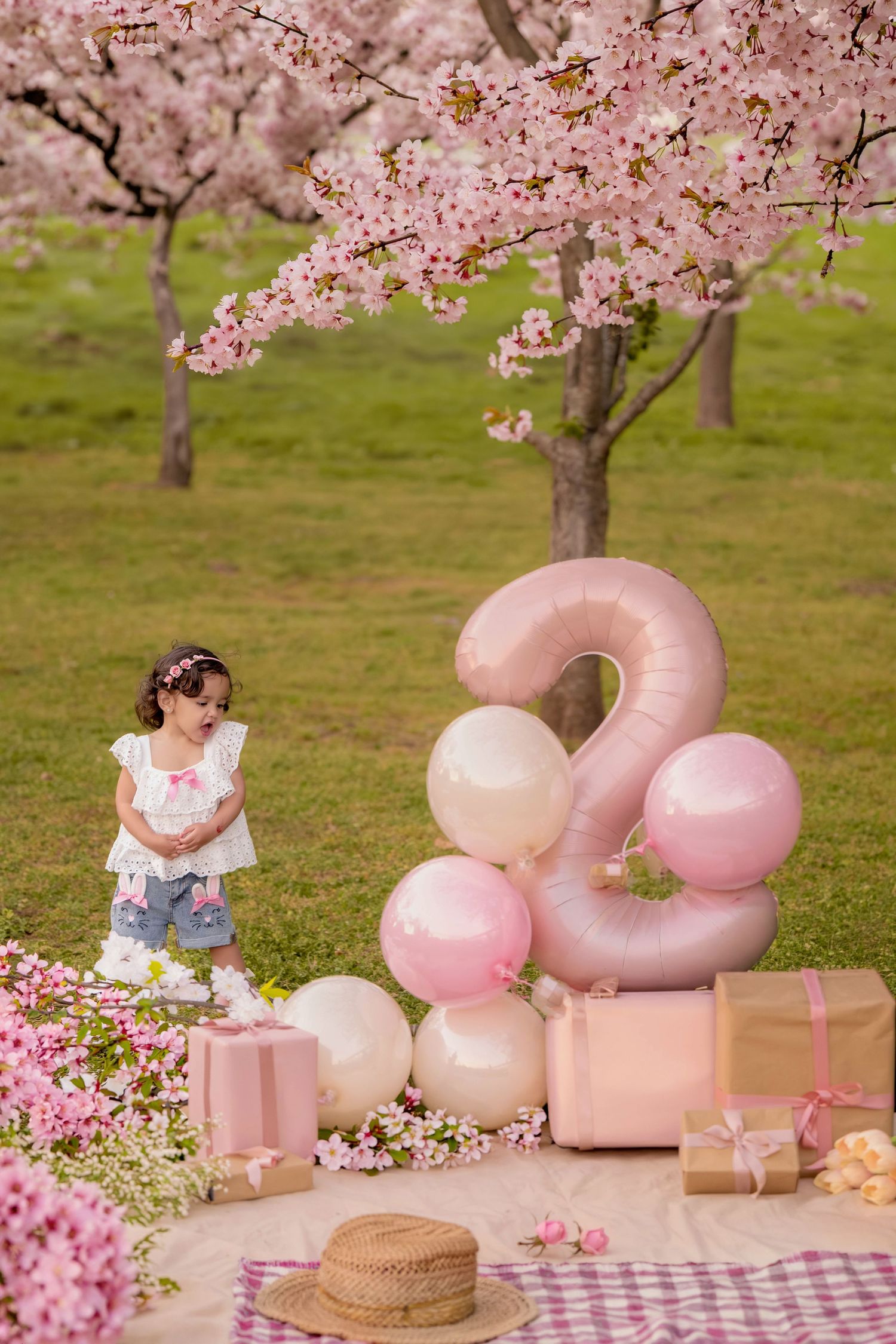 A little girl celebrates her 2nd birthday outdoors with pink balloons, gifts, and cherry blossom trees in bloom.