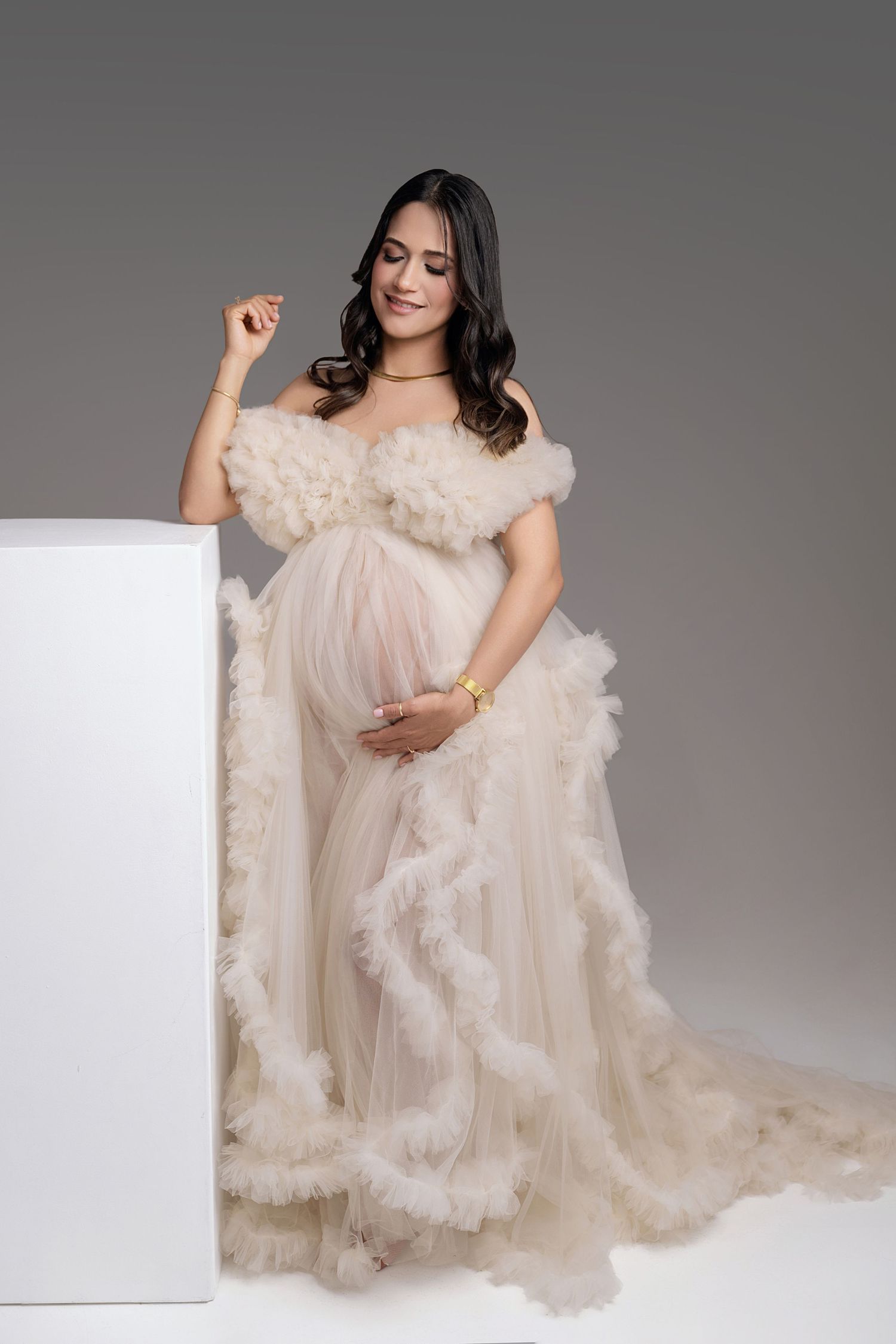 Pregnant woman in elegant cream ruffled off-shoulder tulle maternity gown posing gracefully in studio.