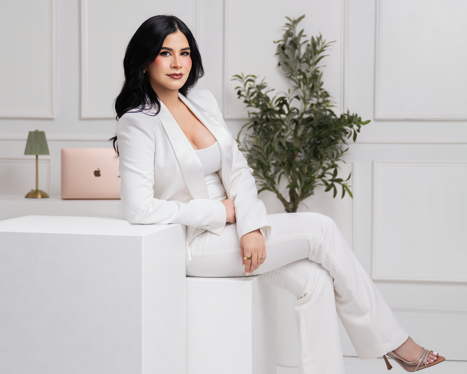 Professional woman in white suit sitting on white cube with laptop, green plant, and gold lamp in bright studio.