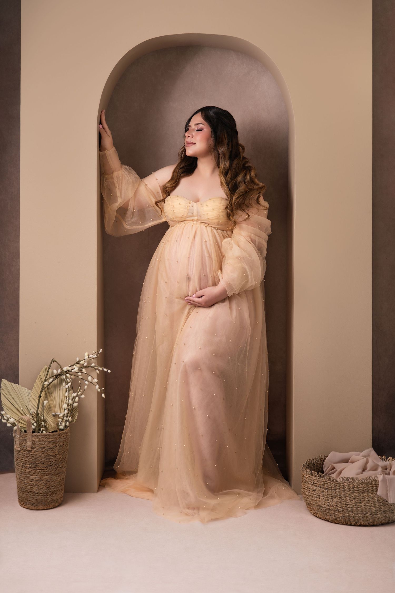 Pregnant woman in elegant sheer peach maternity gown posing in arched alcove with floral decor.