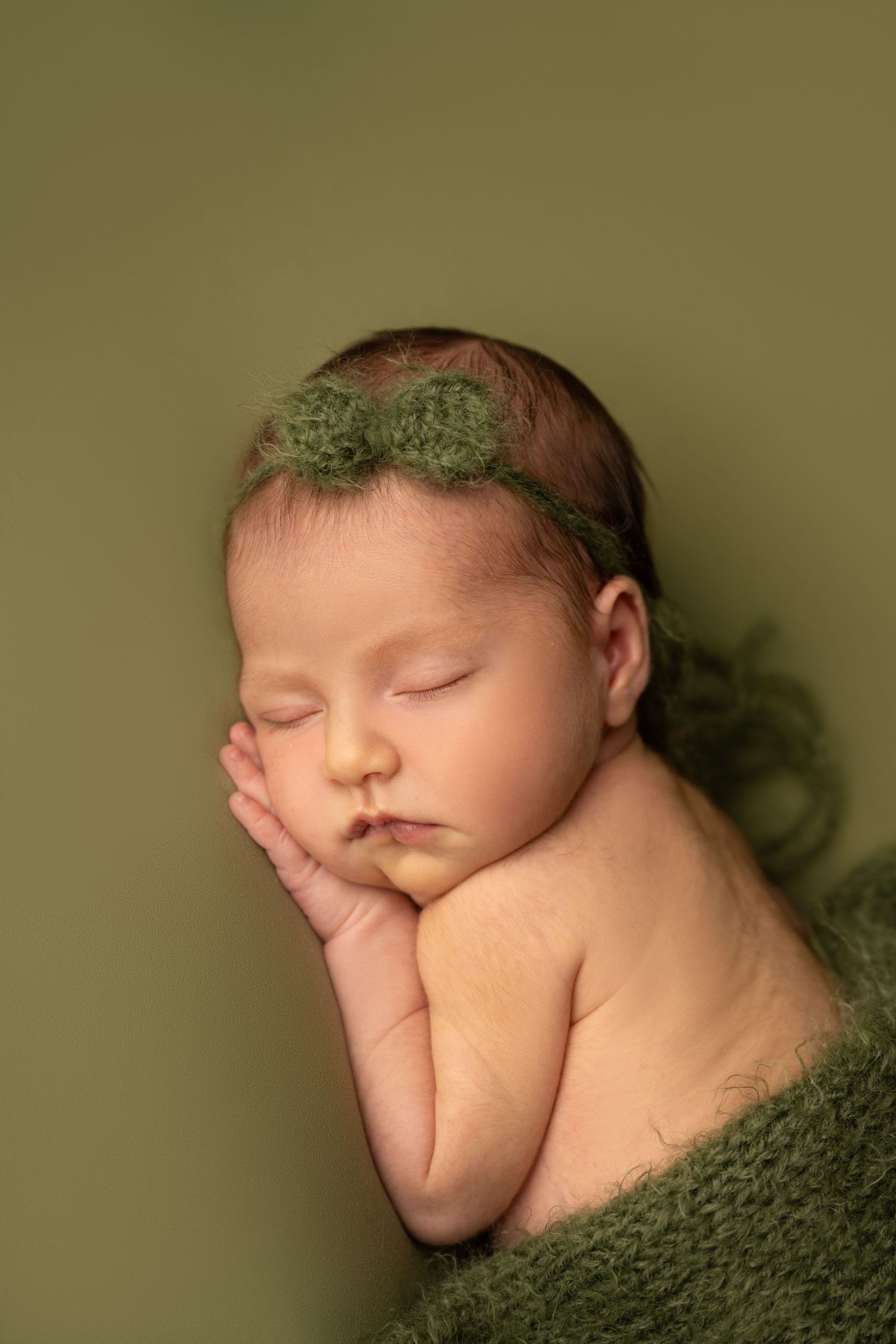 Sleeping newborn wearing green headband against olive background in artistic portrait.