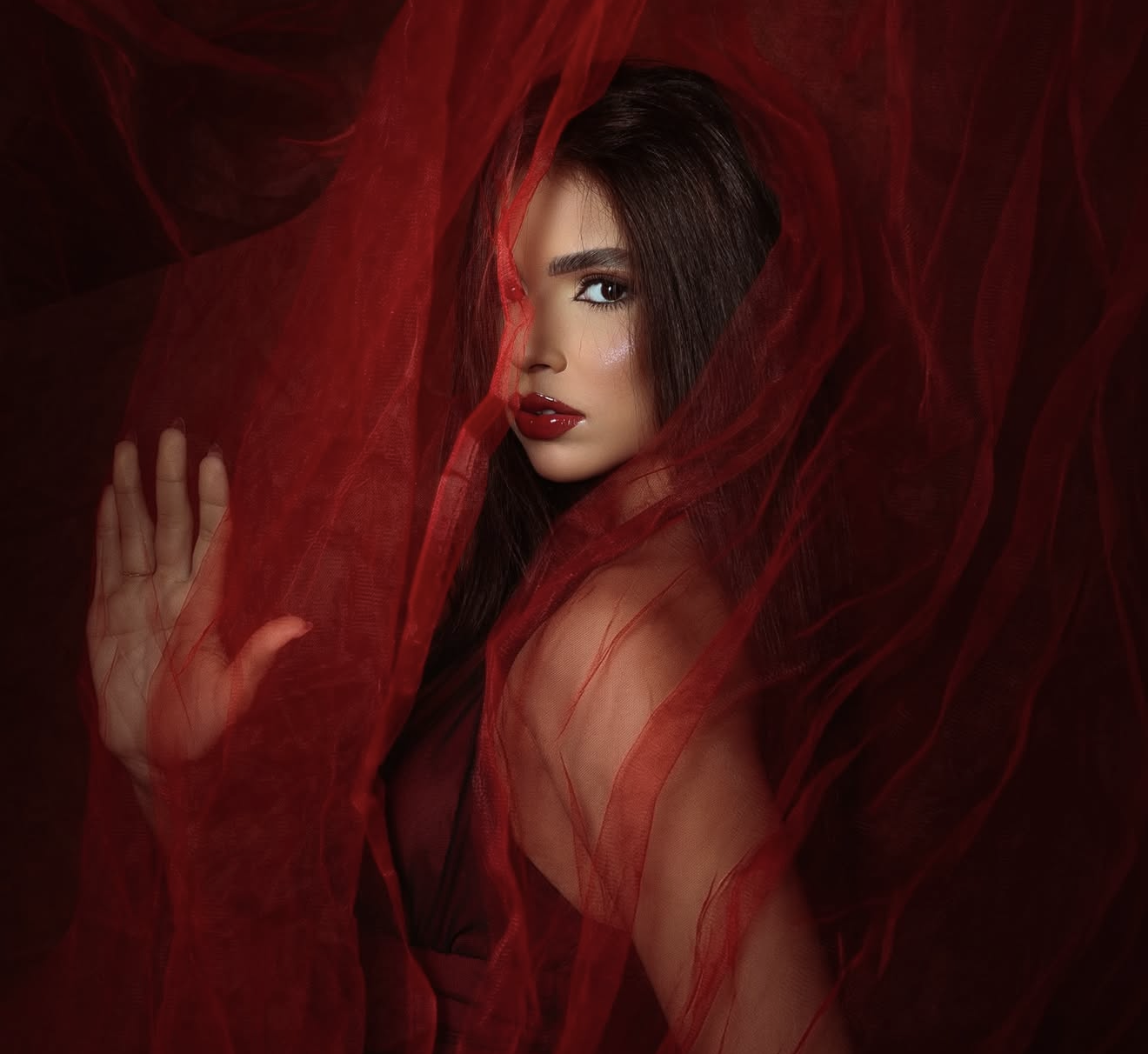 A mysterious woman draped in flowing red fabric with red lips gazes intensely, one hand raised against the sheer cloth.