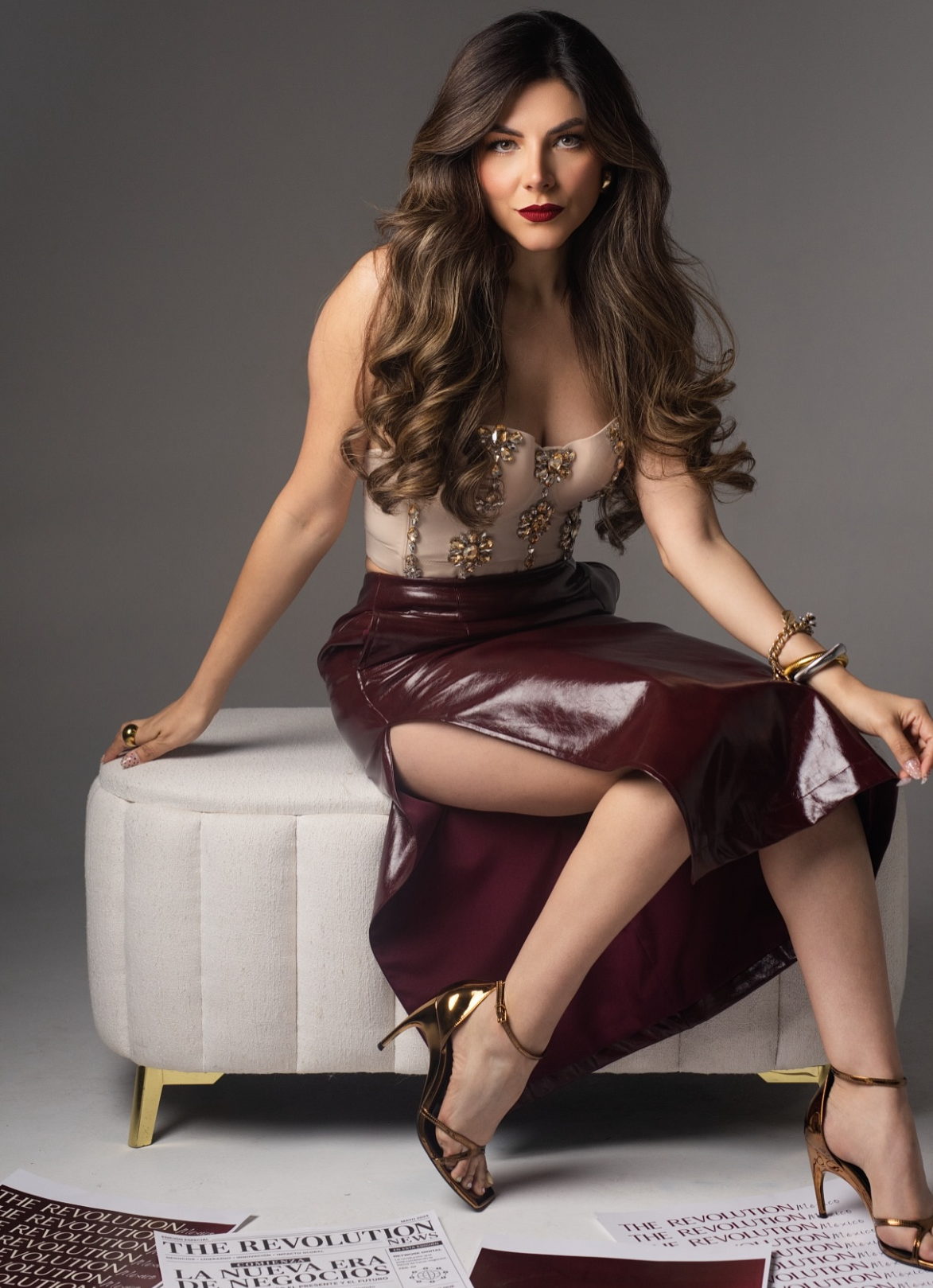 Elegant woman in burgundy leather skirt and embellished top seated on white ottoman with gold heels.