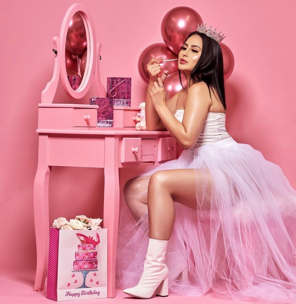 Woman in white tulle skirt and crown sits at pink vanity applying makeup surrounded by pink balloons and birthday gifts.