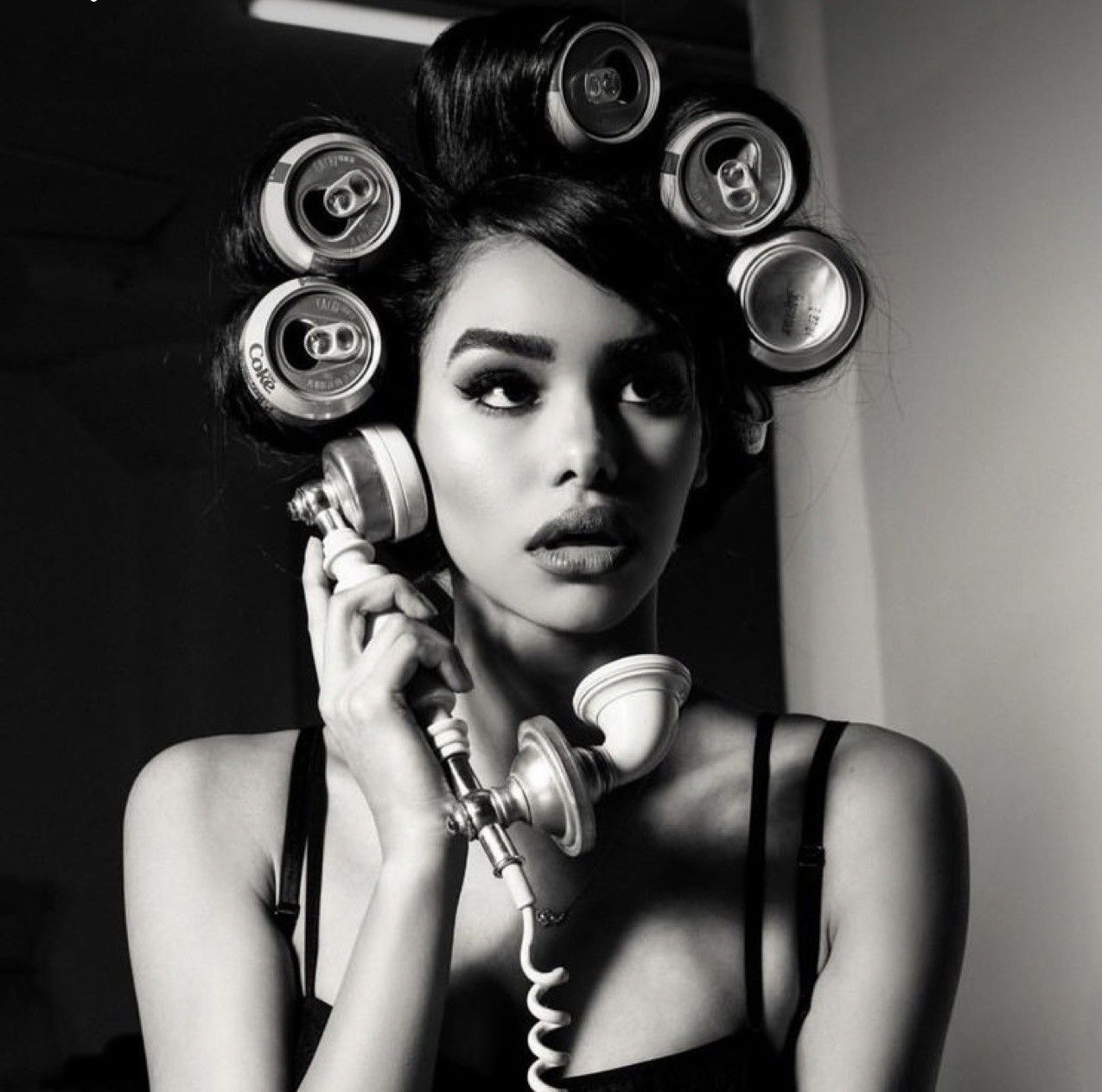 Black and white photo of a woman using a retro phone with soda cans as hair rollers in a glamorous style.