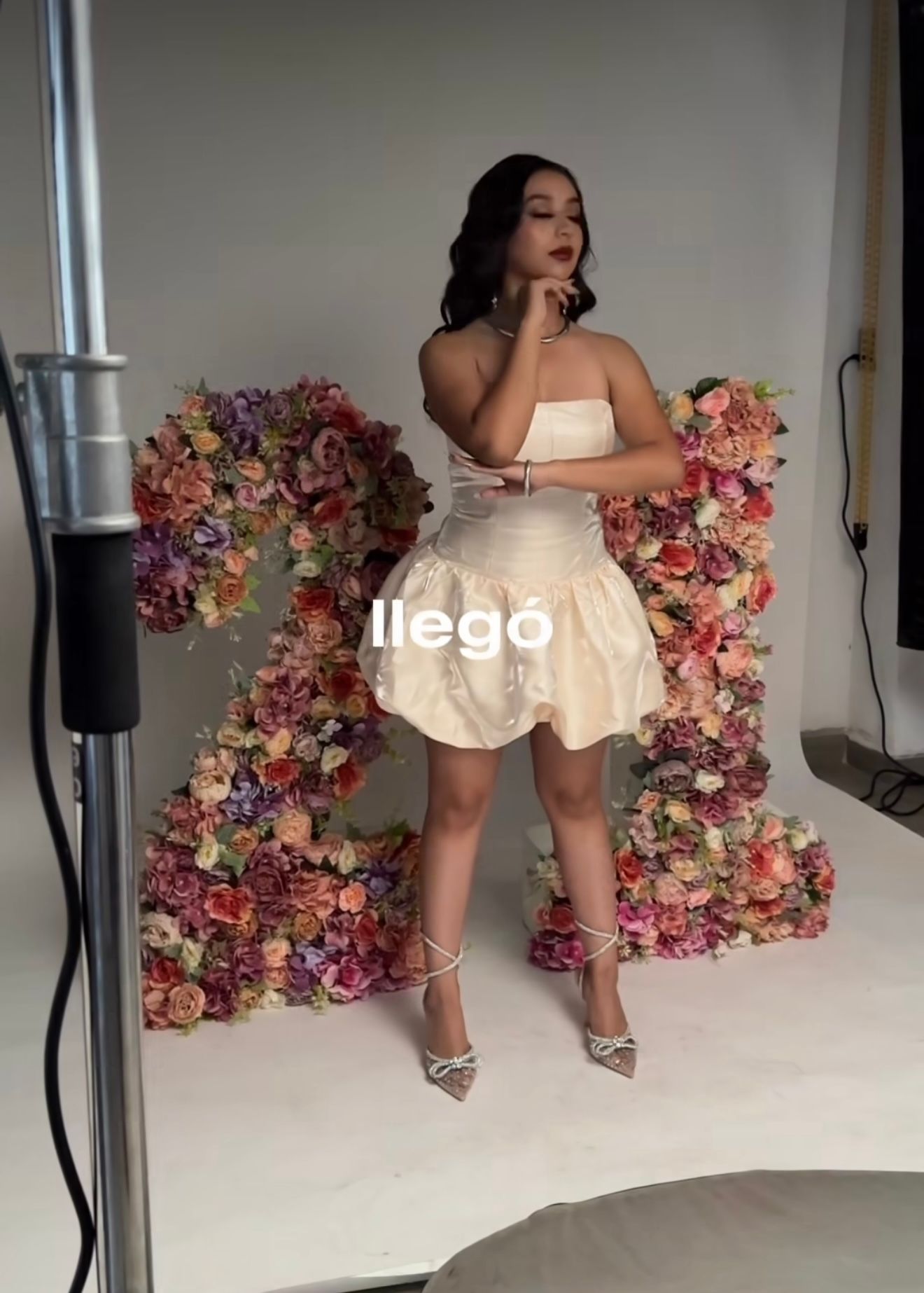 Woman in white strapless bubble mini dress and silver heels poses before colorful floral backdrop with 'llego' text.