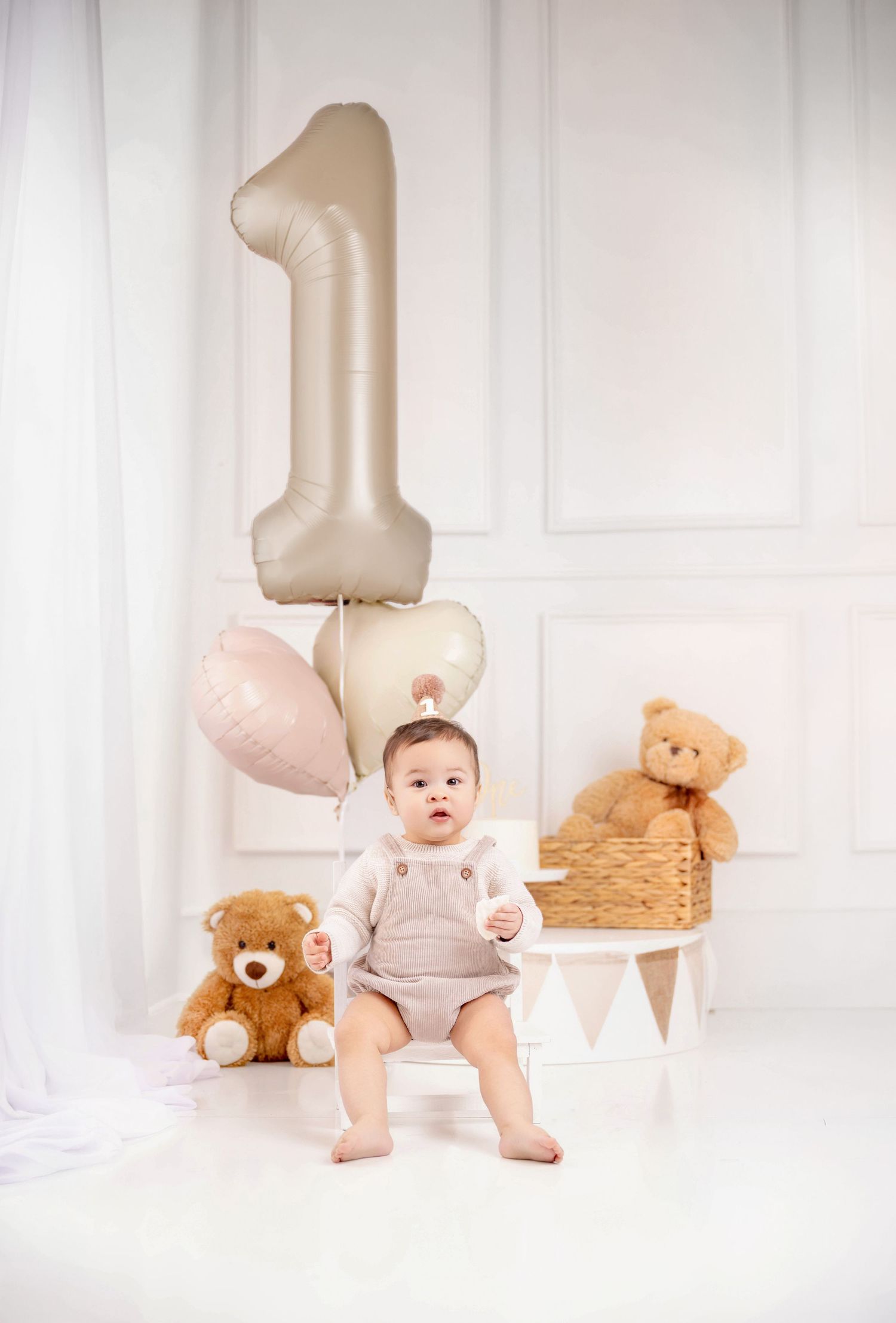 Baby's first birthday portrait with number one balloon, teddy bears, and soft neutral decor in a white studio setting.