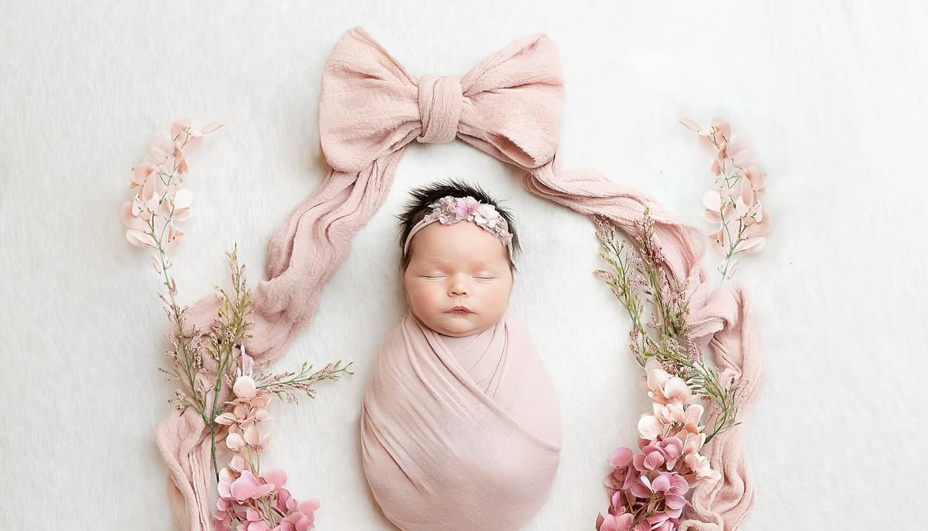 Newborn baby girl swaddled in pink wrap with floral headband, surrounded by pink flowers and bow on white backdrop.