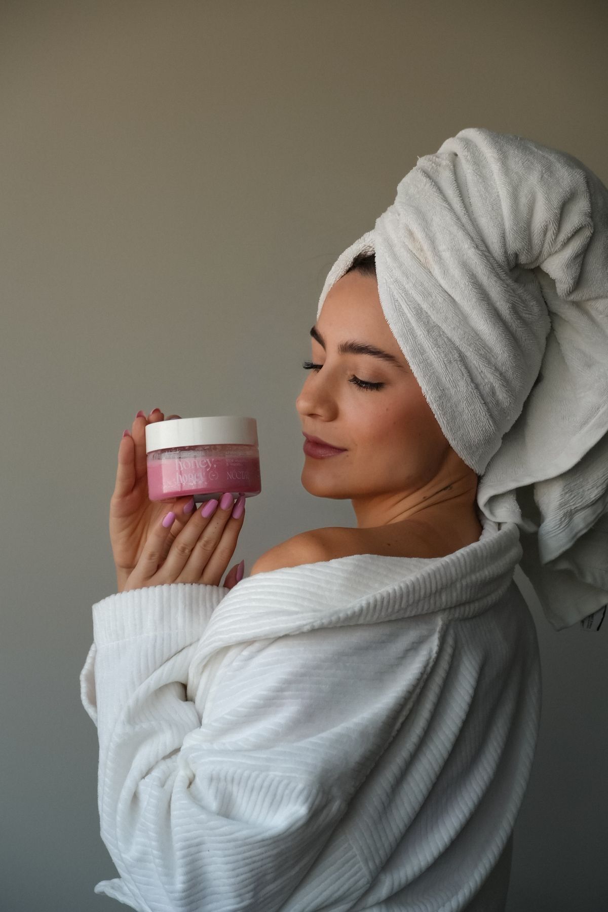 Woman in white bathrobe and towel turban holding pink skincare cream jar with eyes closed.