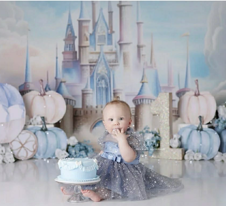 Baby girl in sparkly dress celebrates first birthday with blue cake before a Cinderella castle backdrop.