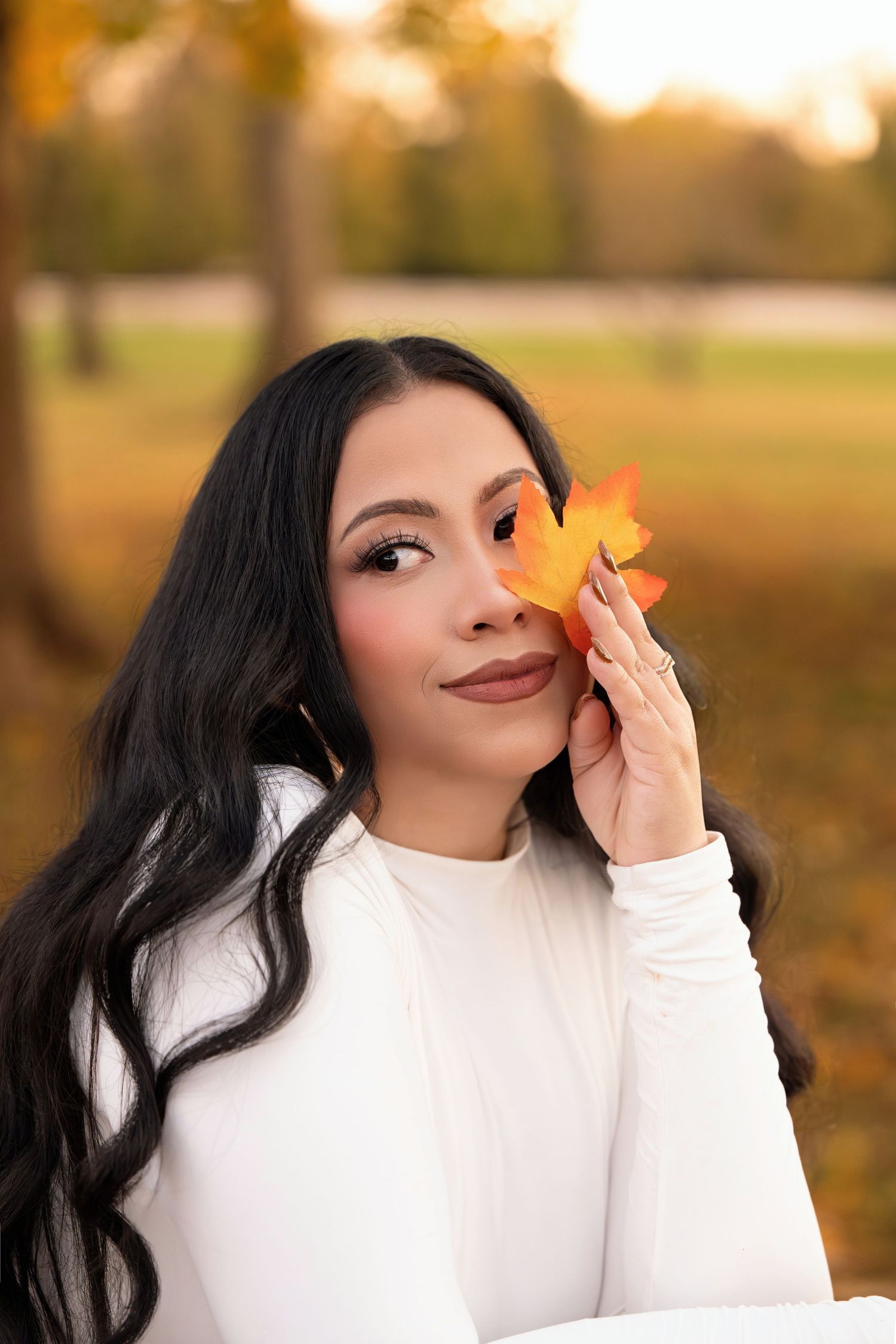 Young woman with long black hair holds an orange maple leaf near her face outdoors in autumn park setting.