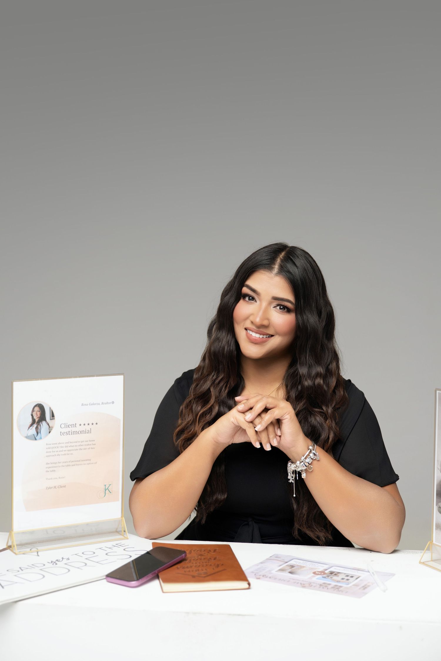 Professional business portrait of someone in black attire smiling while leaning over desk with papers and laptop.