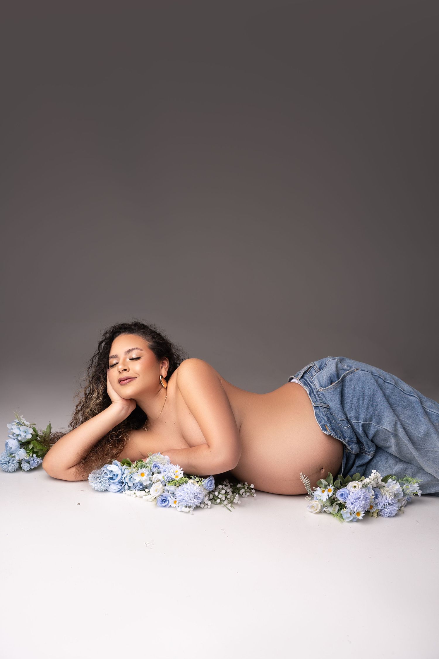 Artistic maternity photo with blue hydrangea flowers scattered around a reclining figure in denim against grey backdrop.