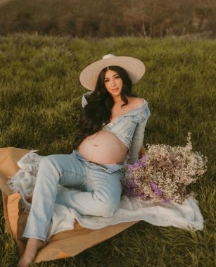 Outdoor maternity photoshoot in meadow with flowers, featuring subject in light blue dress reclining on grass.