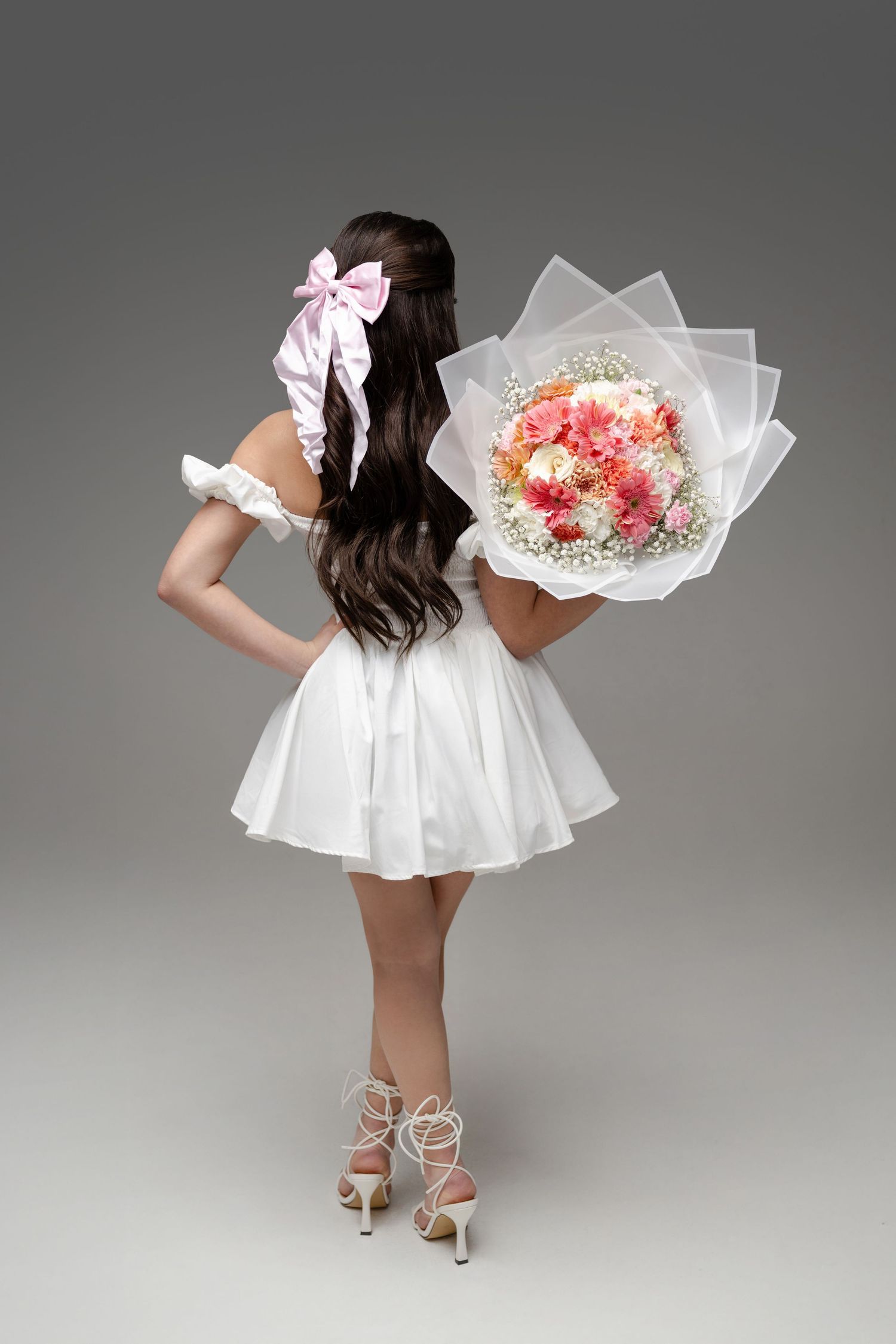 Woman in white mini dress holding pink floral bouquet, viewed from behind, with bow hair accessory and strappy heels.