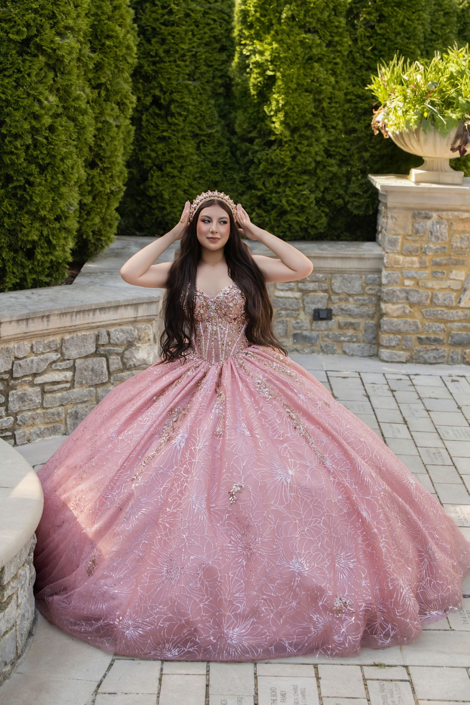 Young woman in a pink glittery ball gown with gold floral details adjusts her tiara in an elegant garden setting.