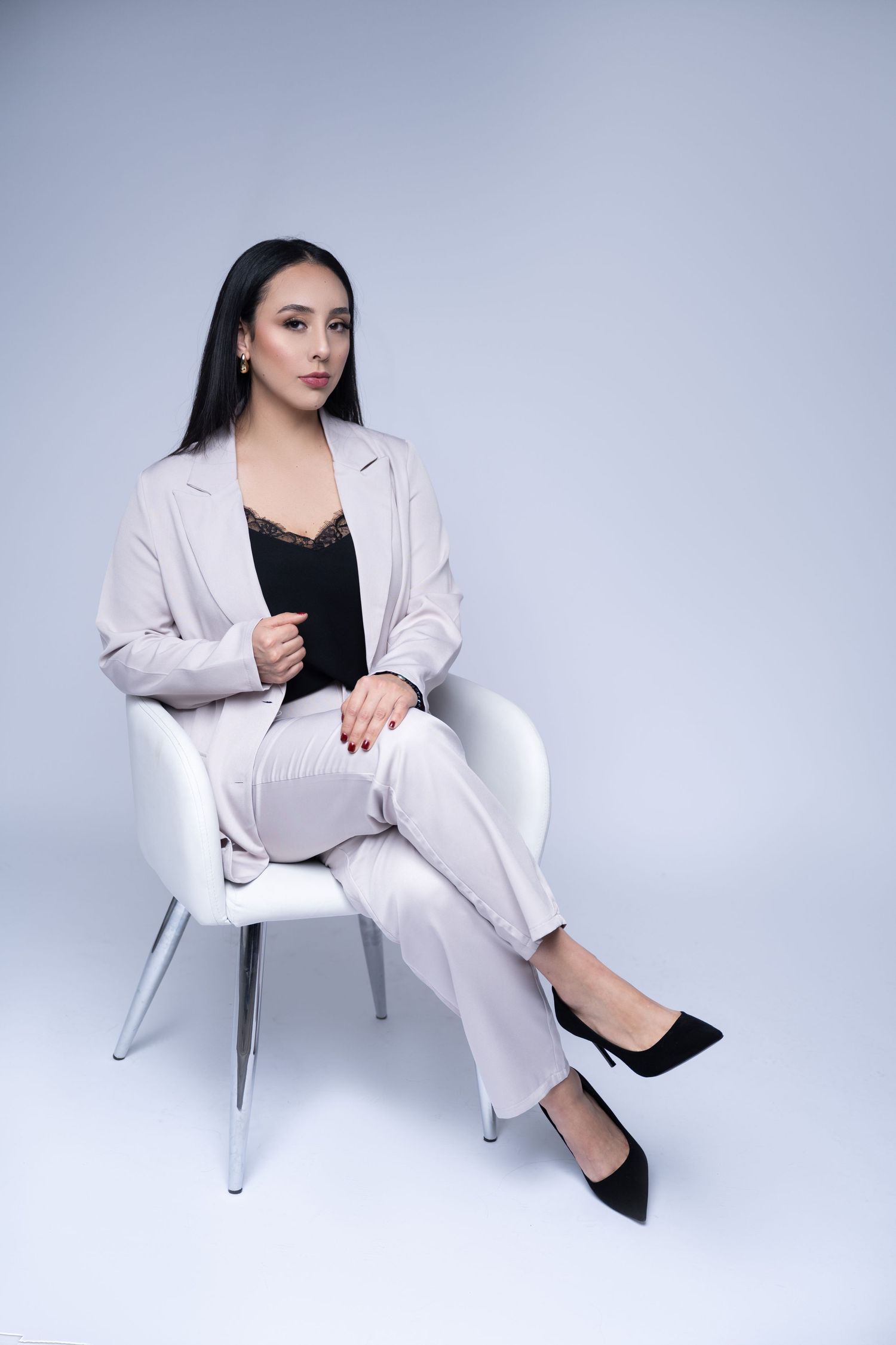 Professional fashion photo of a model in a light gray pantsuit seated elegantly in a white modern chair.