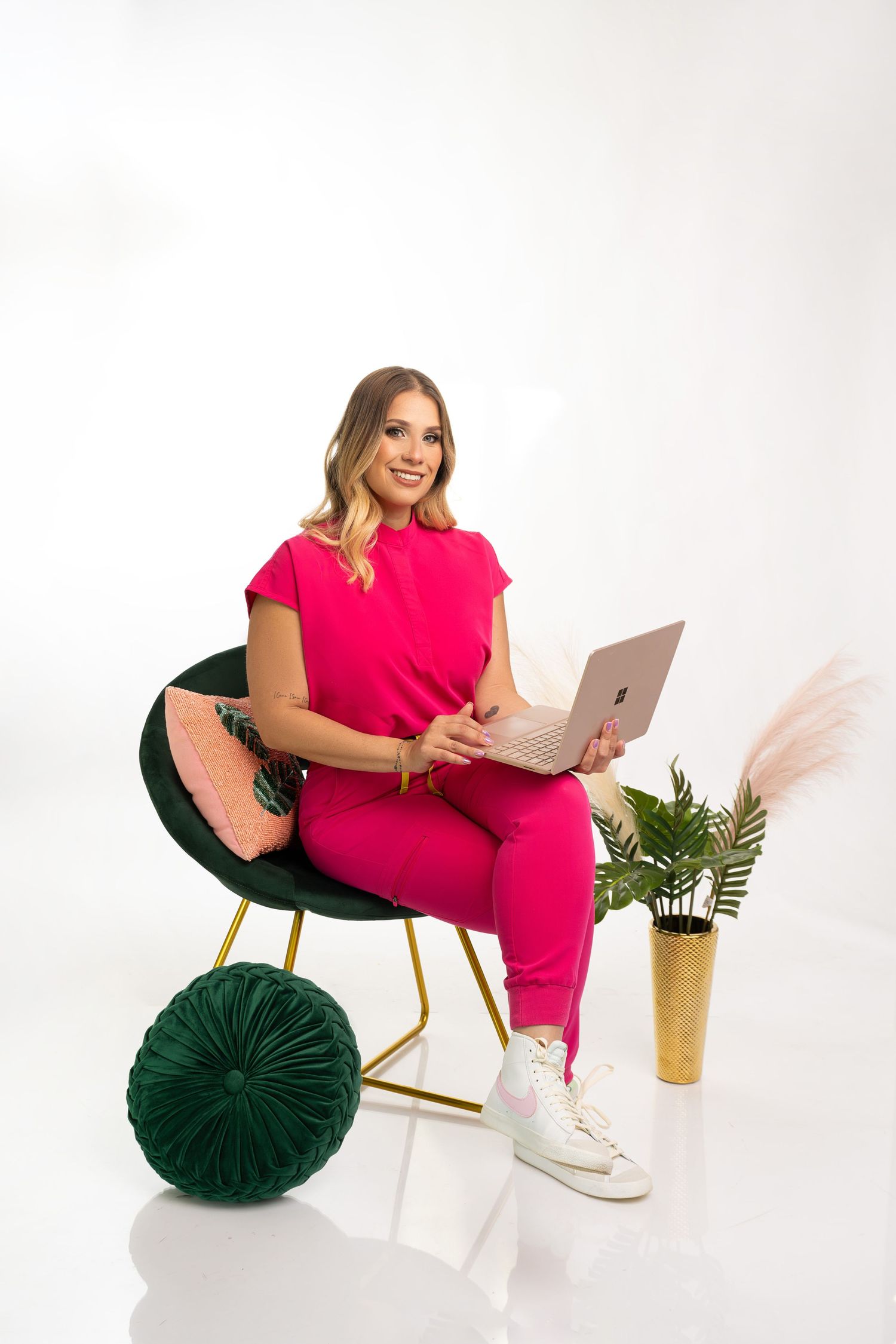 Woman in bright pink outfit working on laptop in modern chair with decorative pillows and plants.