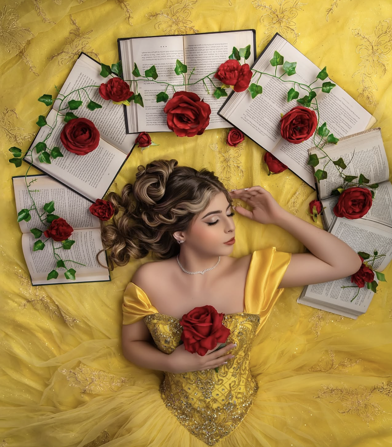 Young woman in yellow Belle gown lying among open books and red roses in a Beauty and the Beast inspired portrait.