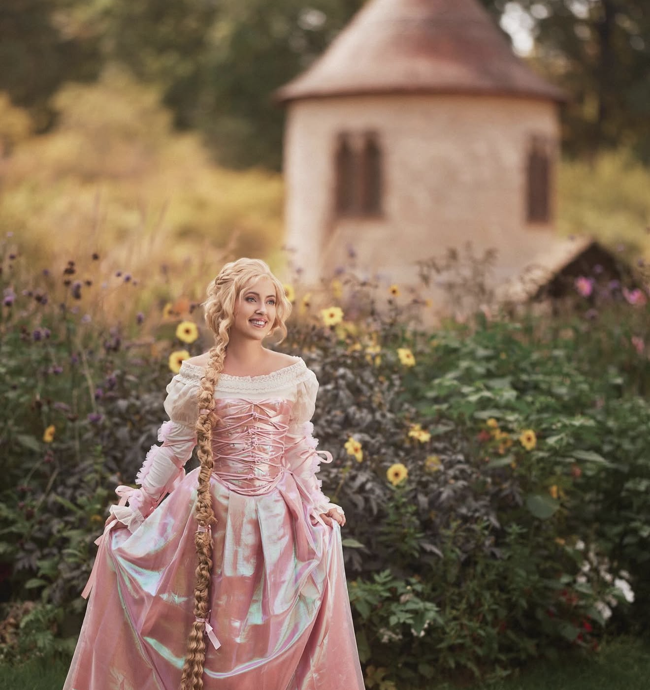 Woman in pink princess gown with long braid poses in garden near stone tower, cosplaying as Rapunzel.