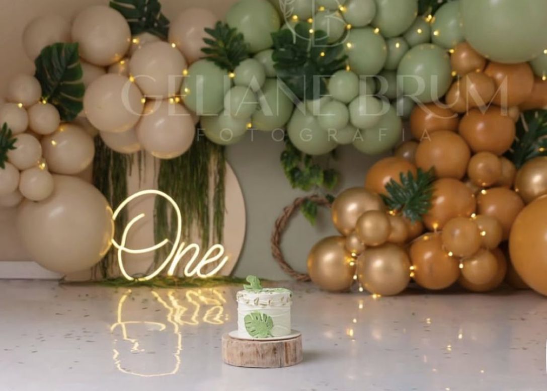 First birthday party setup with elegant balloon arch in peach, sage green and gold tones with illuminated 'One' neon sign.