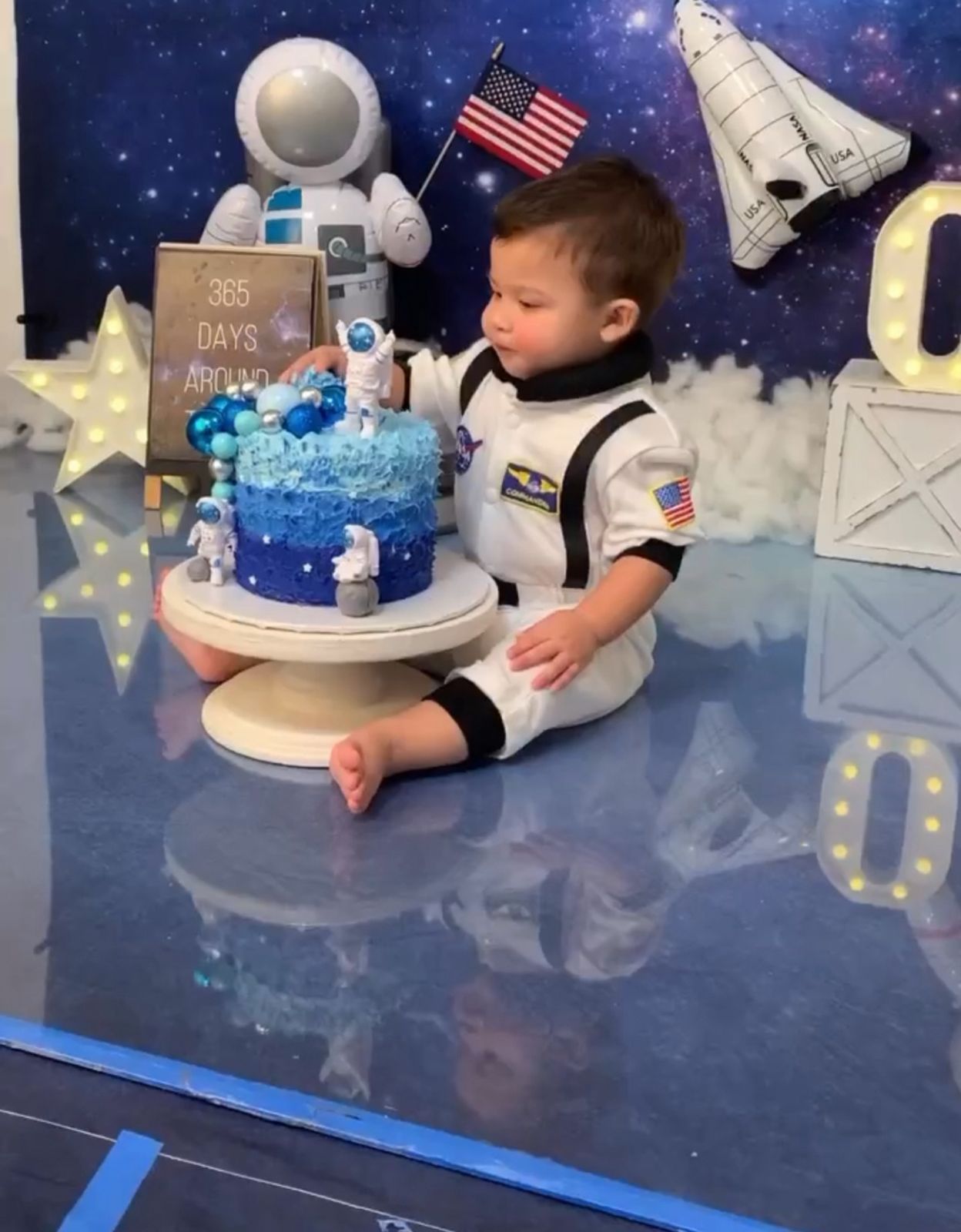 A space-themed birthday party setup with a blue cake, astronaut decoration, American flag, and space shuttle against a starry backdrop.