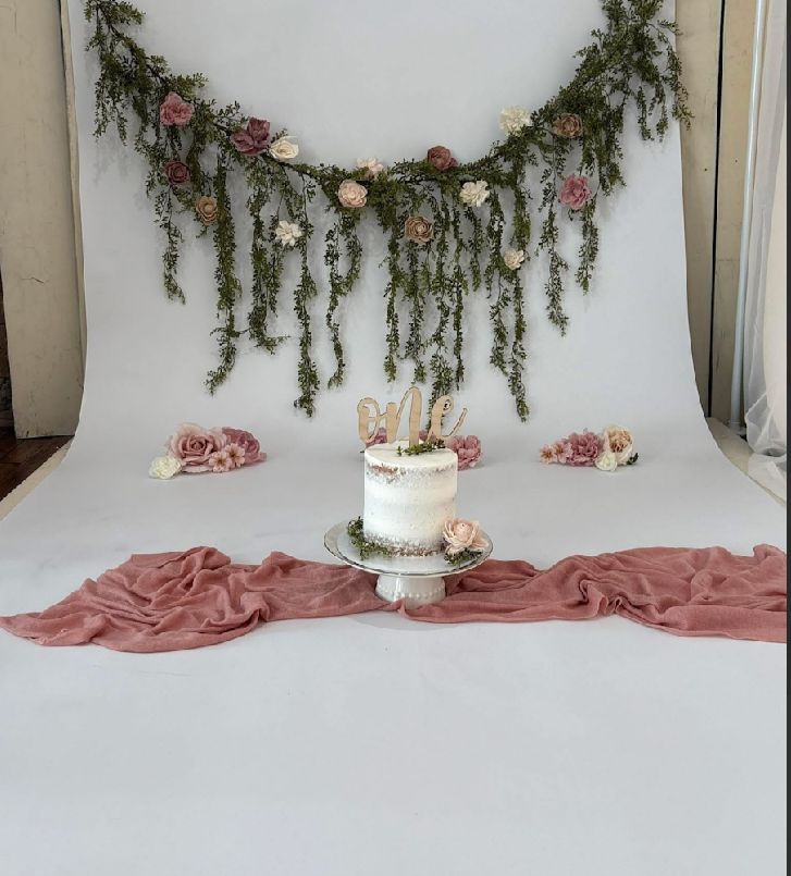 A rustic wedding cake display with dried floral garland backdrop, pink fabric runner, and white tiered cake on a stand.